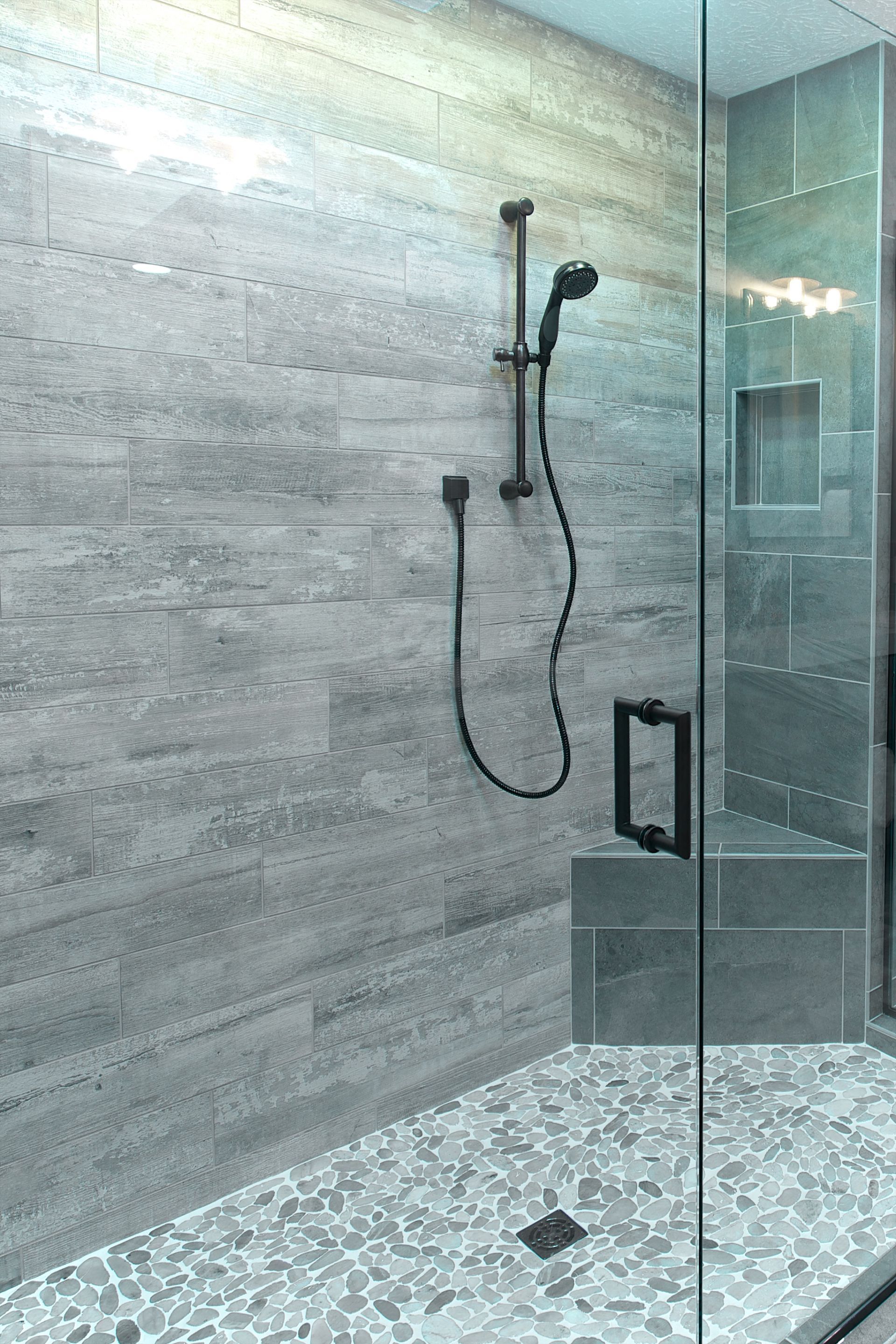 Gray tiled shower with glass door, handheld showerhead, pebble floor, and recessed shelf.