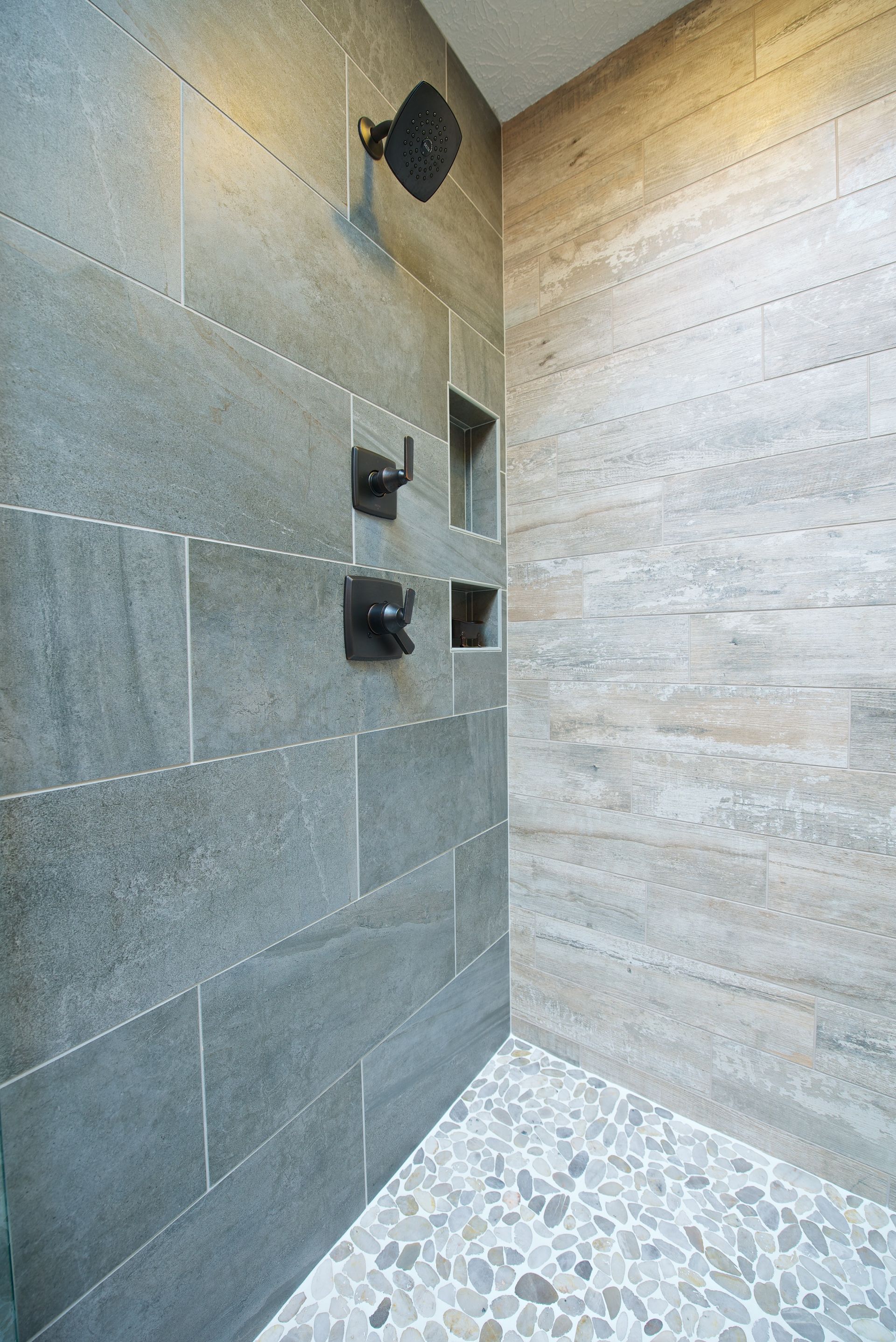 Shower with gray and beige tile walls, pebble floor, and dark fixtures.