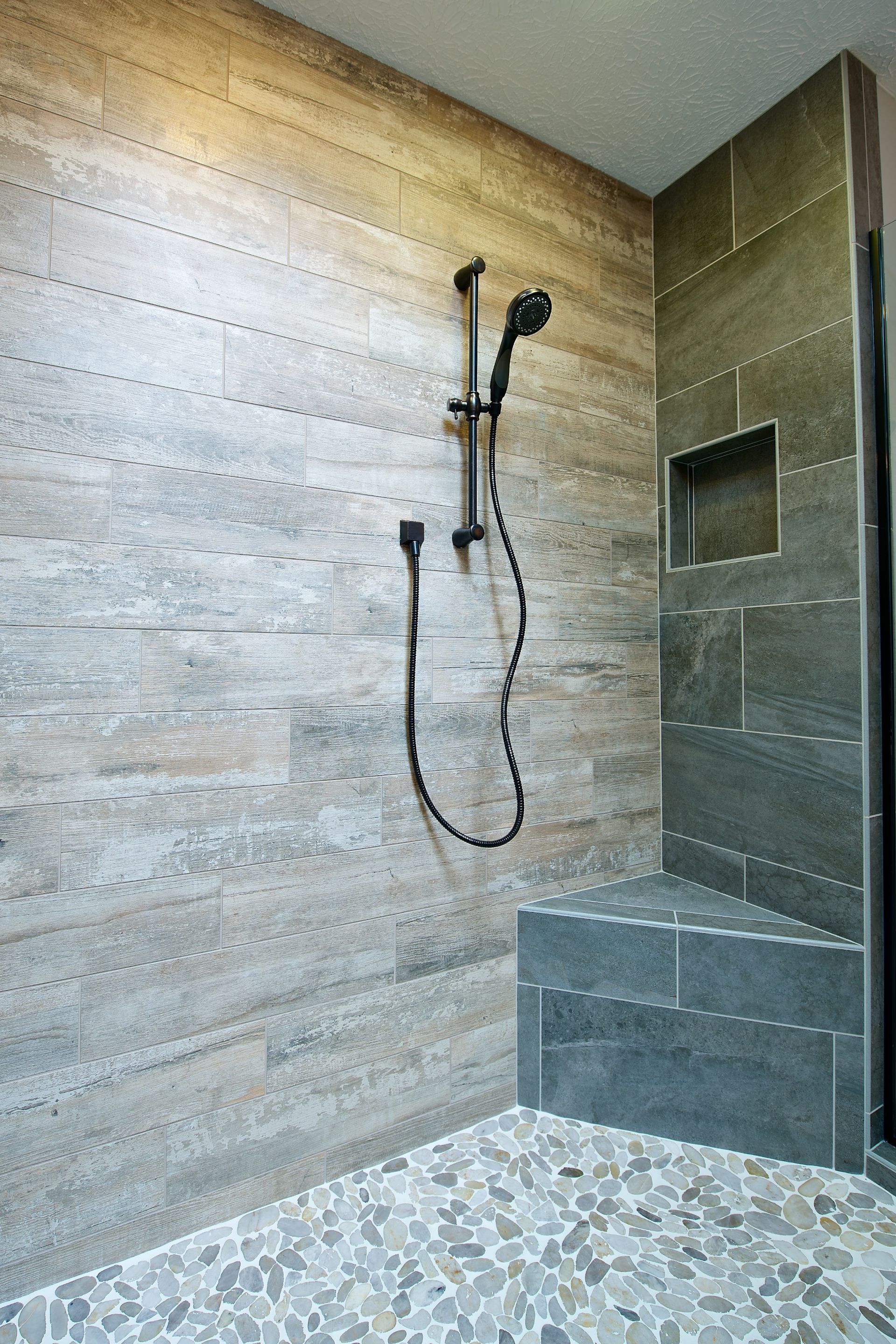 Shower with gray and wood-look tile, pebble floor, built-in seat, and black shower fixture.