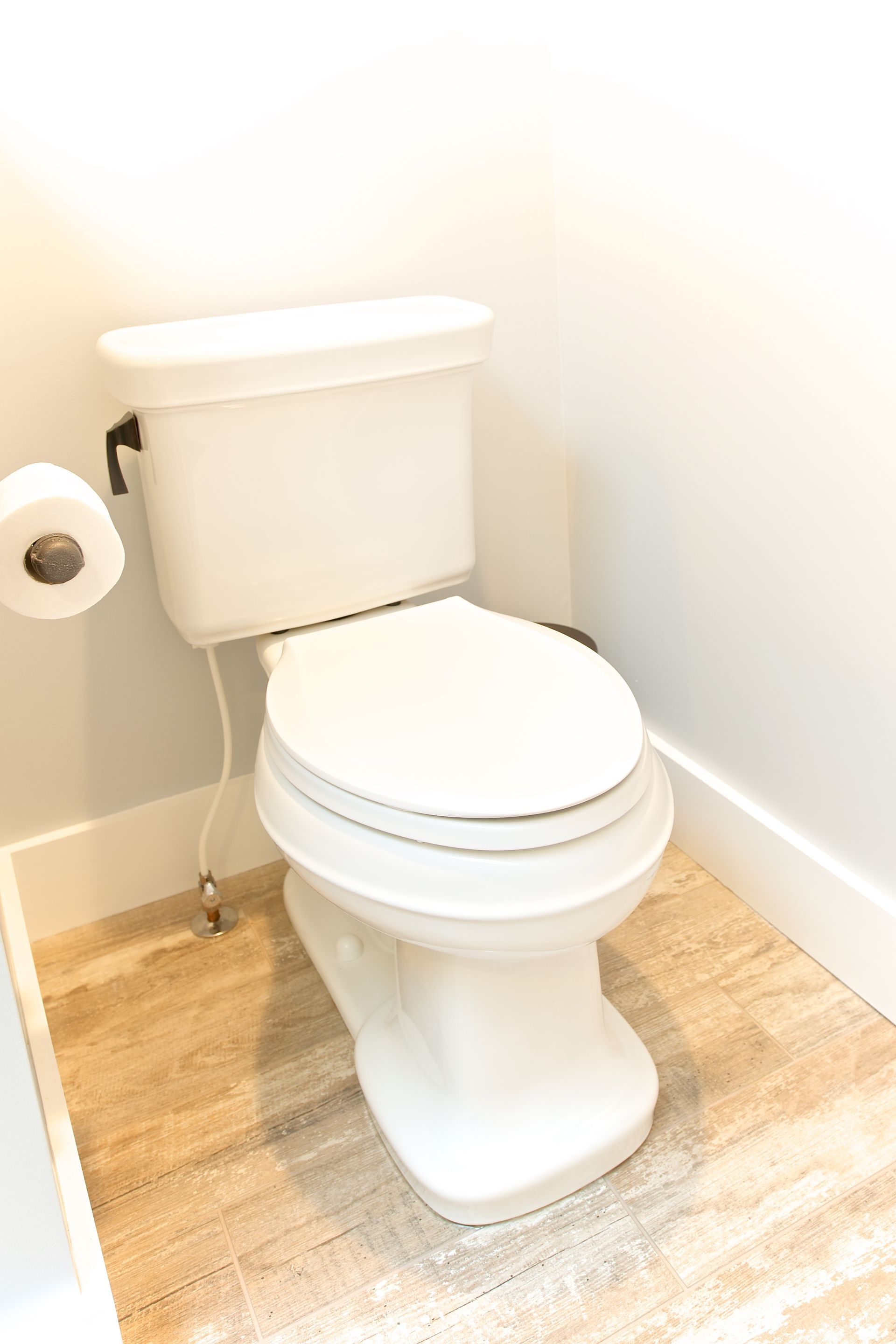 White toilet in a bathroom with wood-look flooring and a roll of toilet paper on the left.