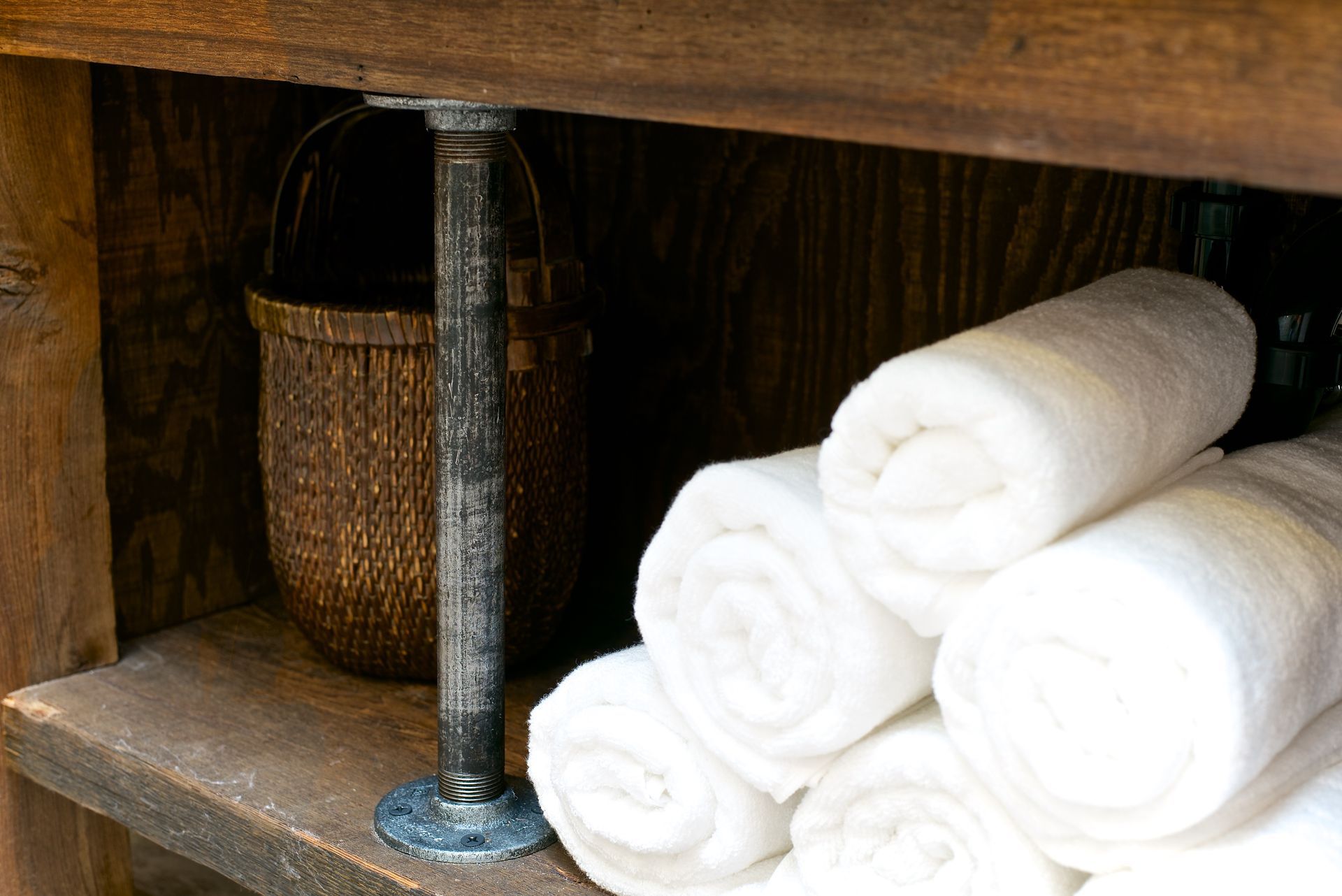 White rolled towels on wooden shelf with a basket, supported by a metal pipe.