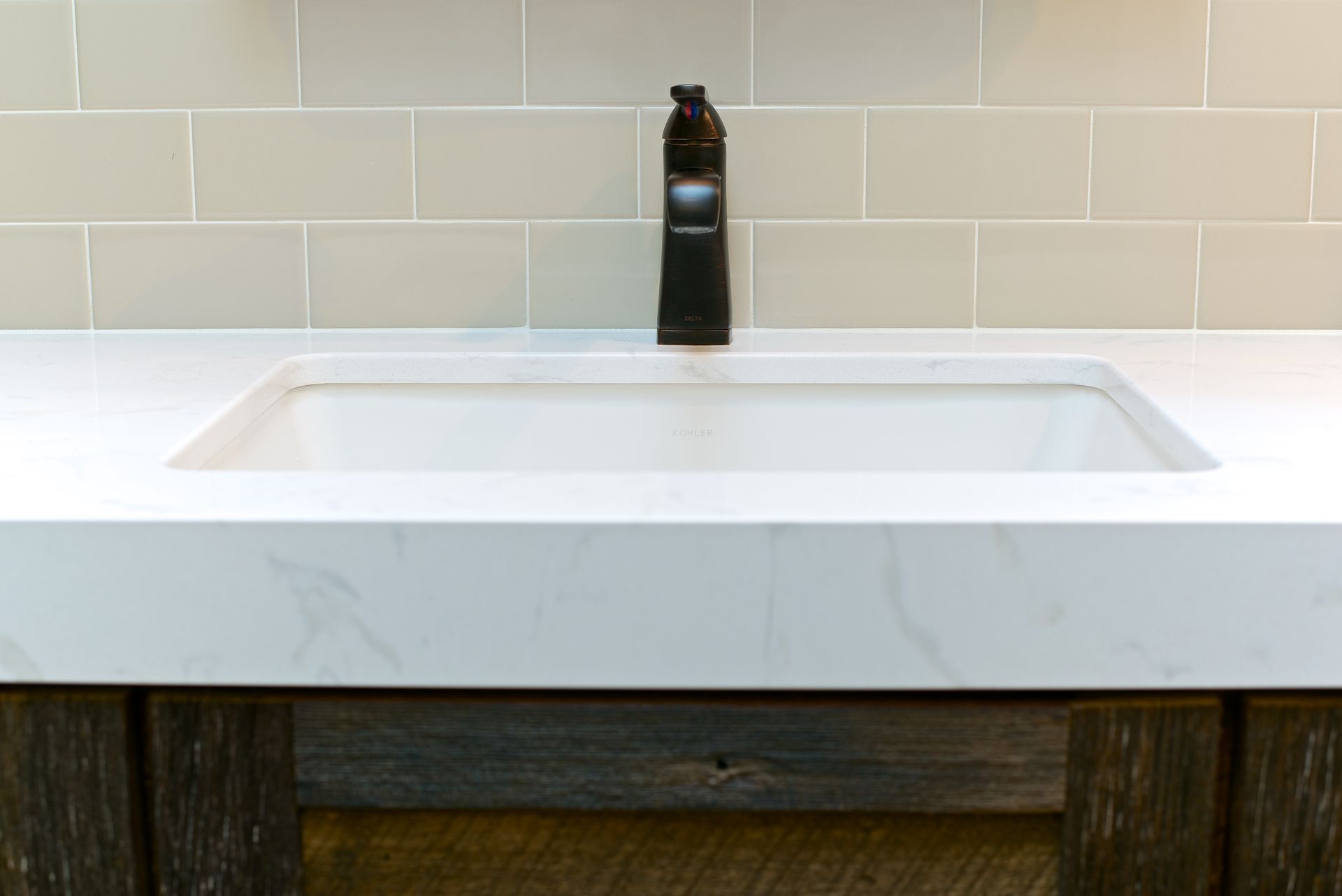 White sink and dark faucet on a light-colored countertop. Dark wood cabinet below with tiled wall background.