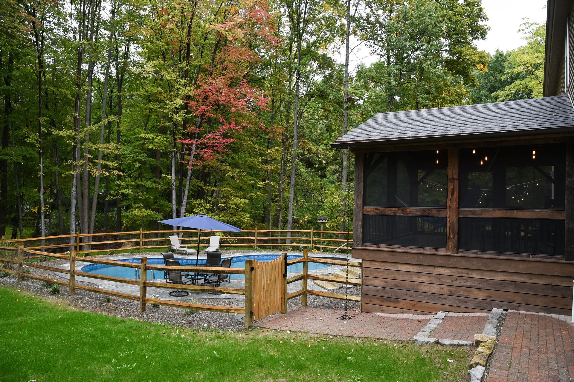 Screened porch next to a pool with a fence, surrounded by a green grassy lawn and trees.