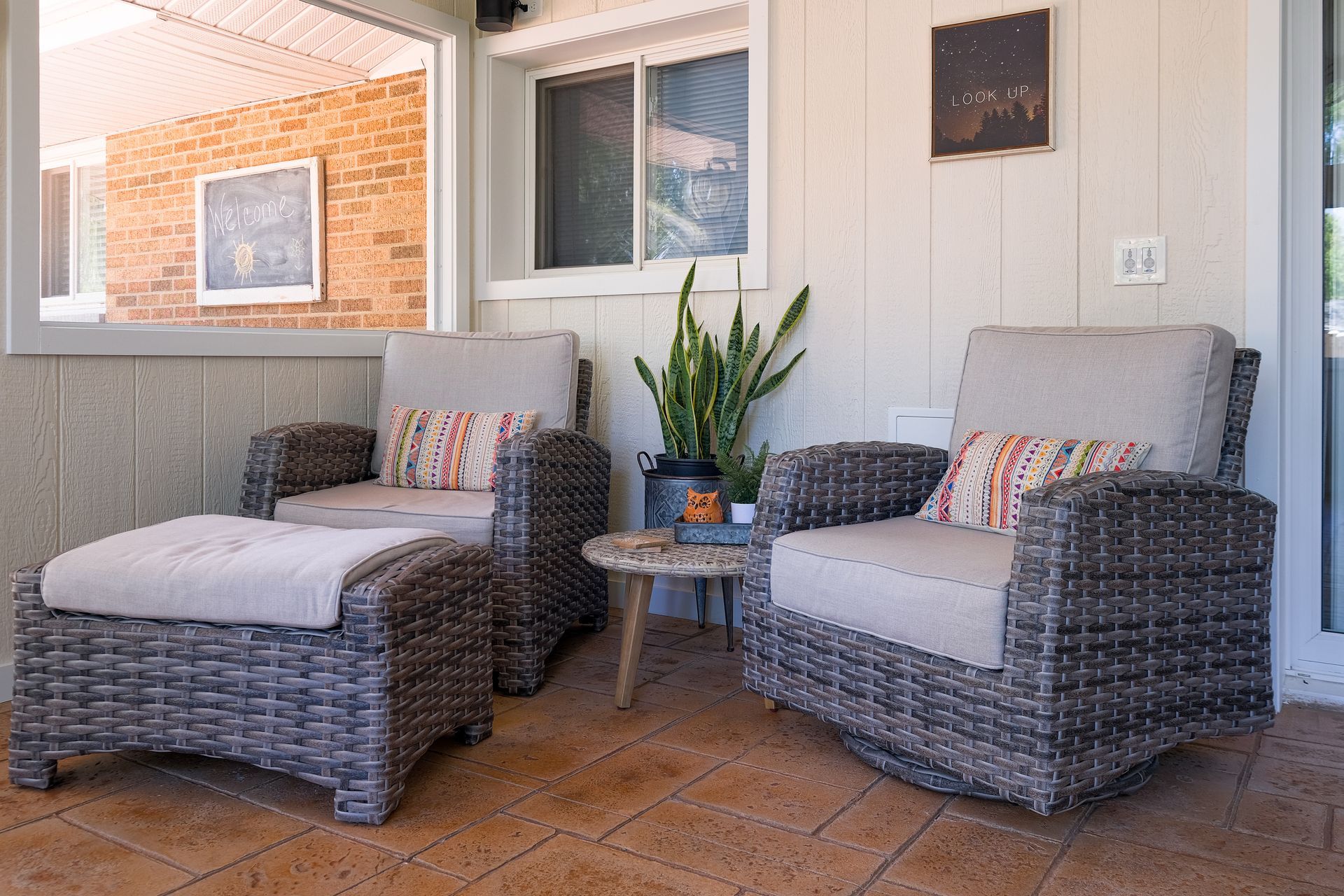 Two wicker chairs with cushions and a footstool on a patio, with a plant and small table.