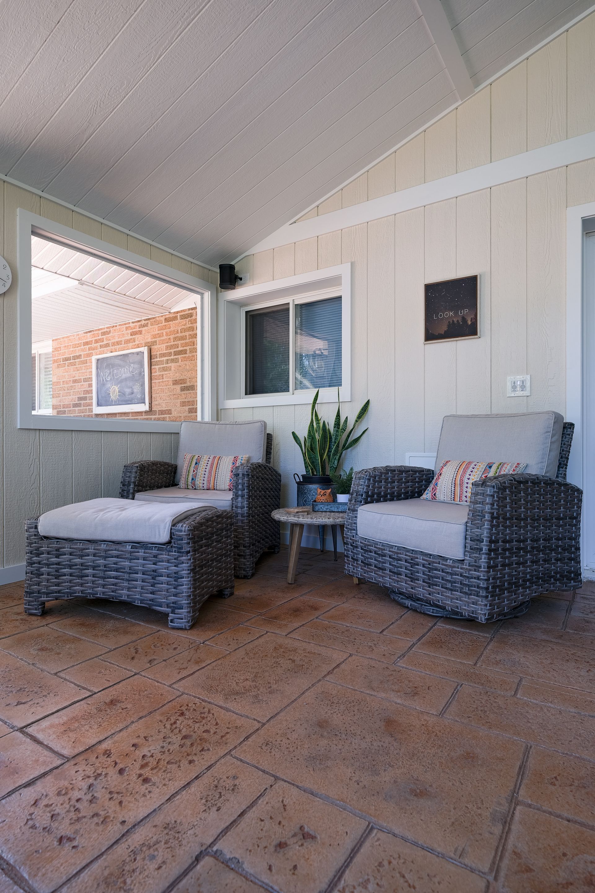 A sunroom with wicker chairs, a small table, and a window overlooking a brick wall.