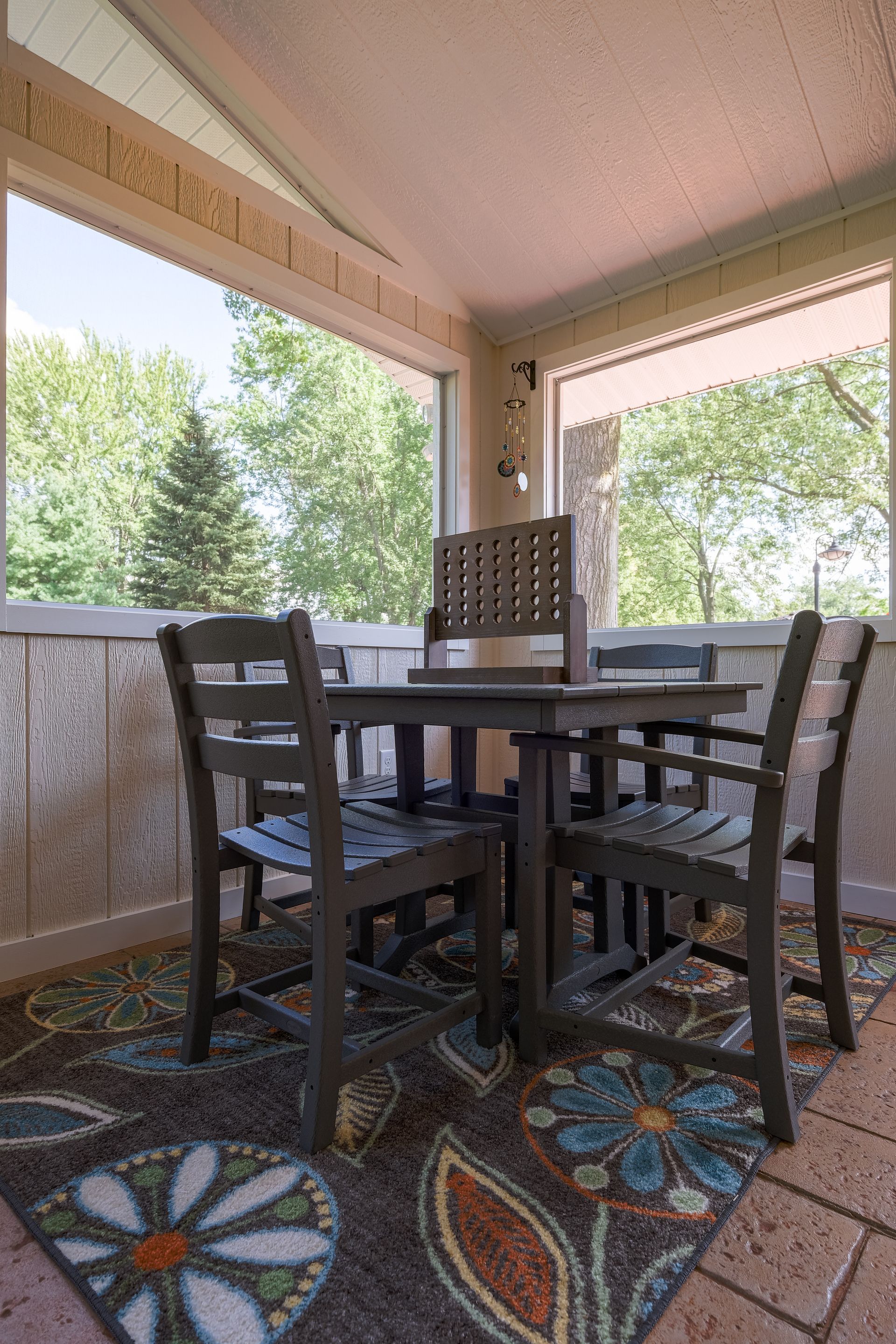 Enclosed patio with a wooden table, chairs, and a floral rug, overlooking a lush green treeline.