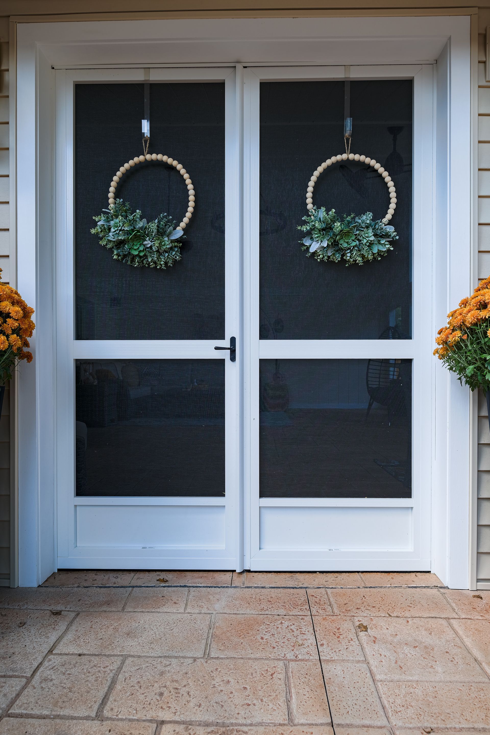White double screen doors with wreaths, framed by siding, and flanked by orange flowers in pots.