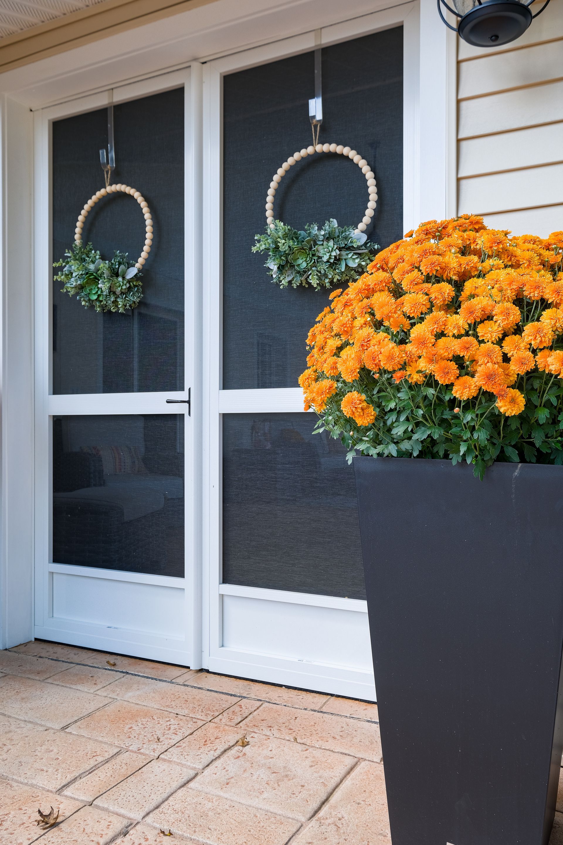 Two wreaths on a screen door, orange mums in a black planter on a brick porch.
