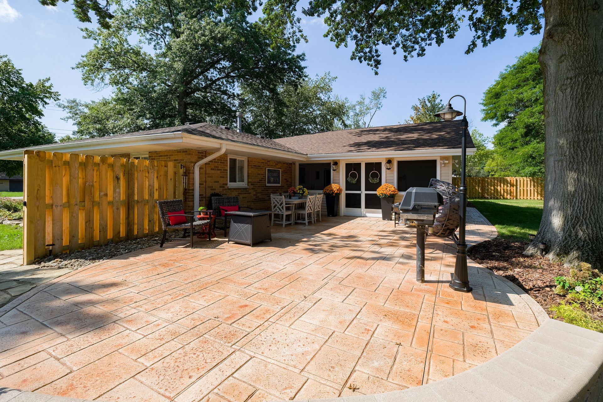 Backyard patio with wooden fence, brick patio, grill, table, chairs, and trees.