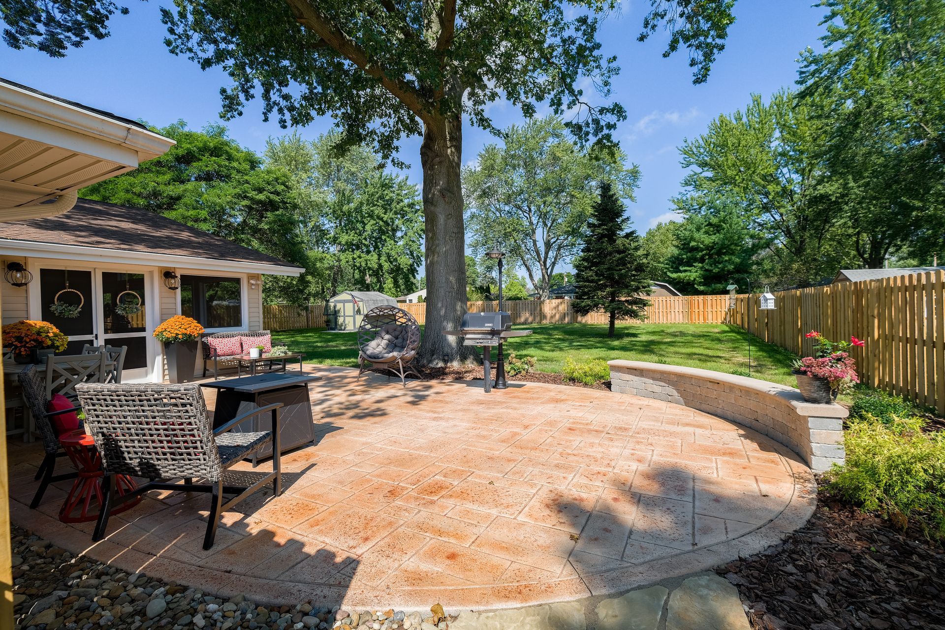 Backyard patio with seating, grill, and stone wall, surrounded by trees and grass. Sunny day.