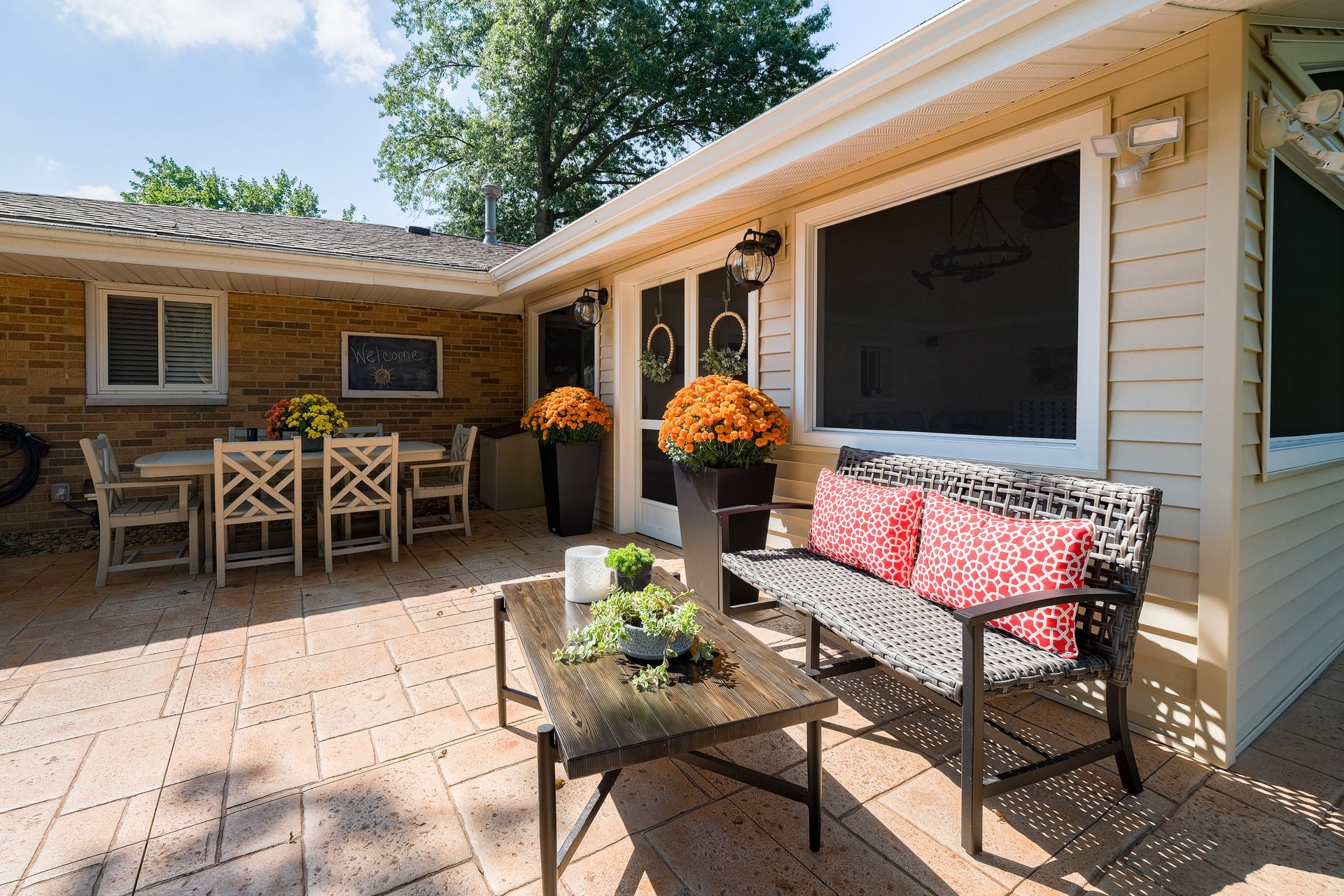 Patio with brick wall, dining table, wicker bench, and potted plants under a sunny sky.