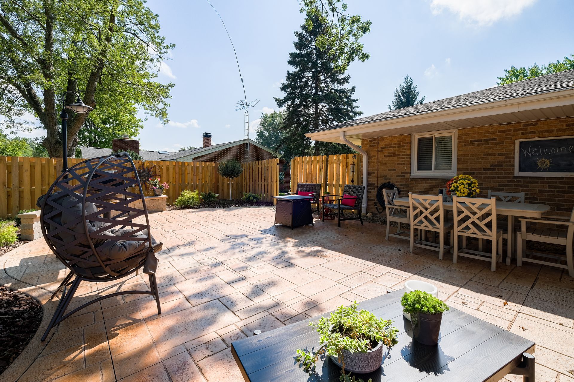Backyard patio with seating, dining table, and hanging chair, surrounded by a wooden fence and greenery.