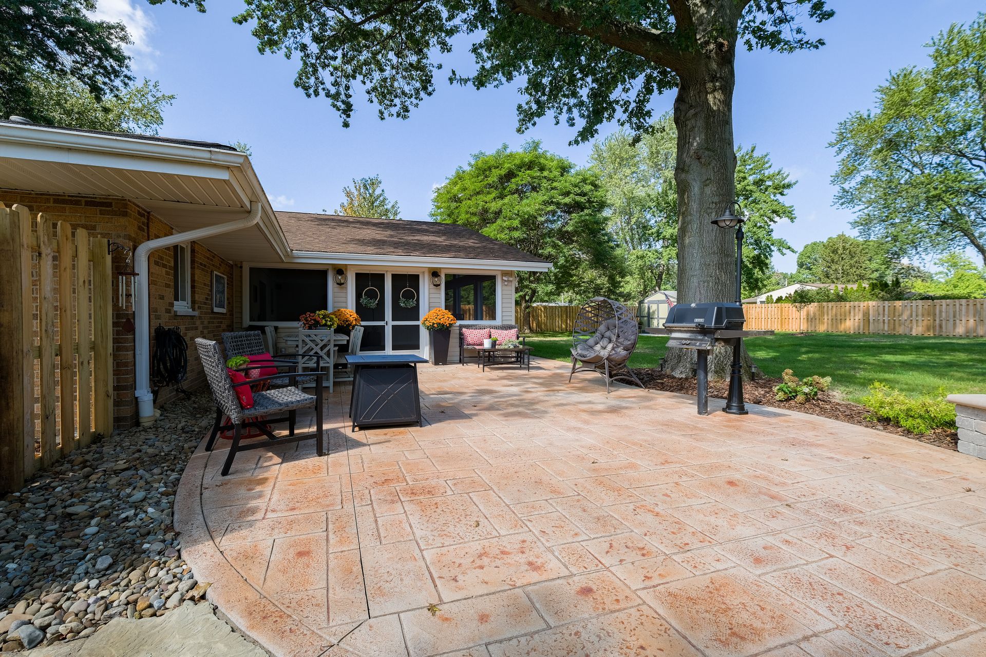 Patio with a grill and chairs next to a house with a wooden fence and large tree, sunny day.