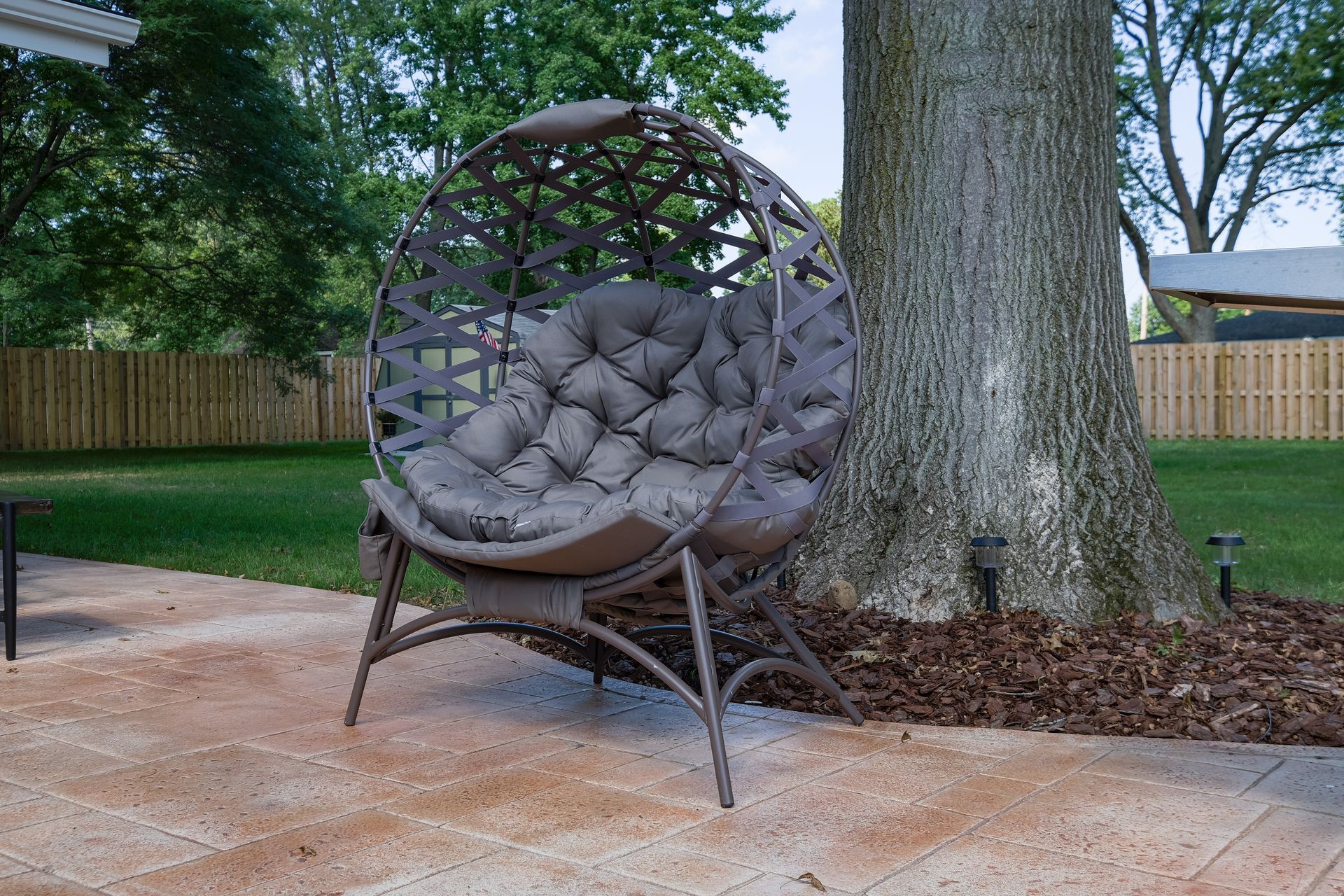 Wicker egg chair with gray cushions on a brick patio next to a tree in a backyard.