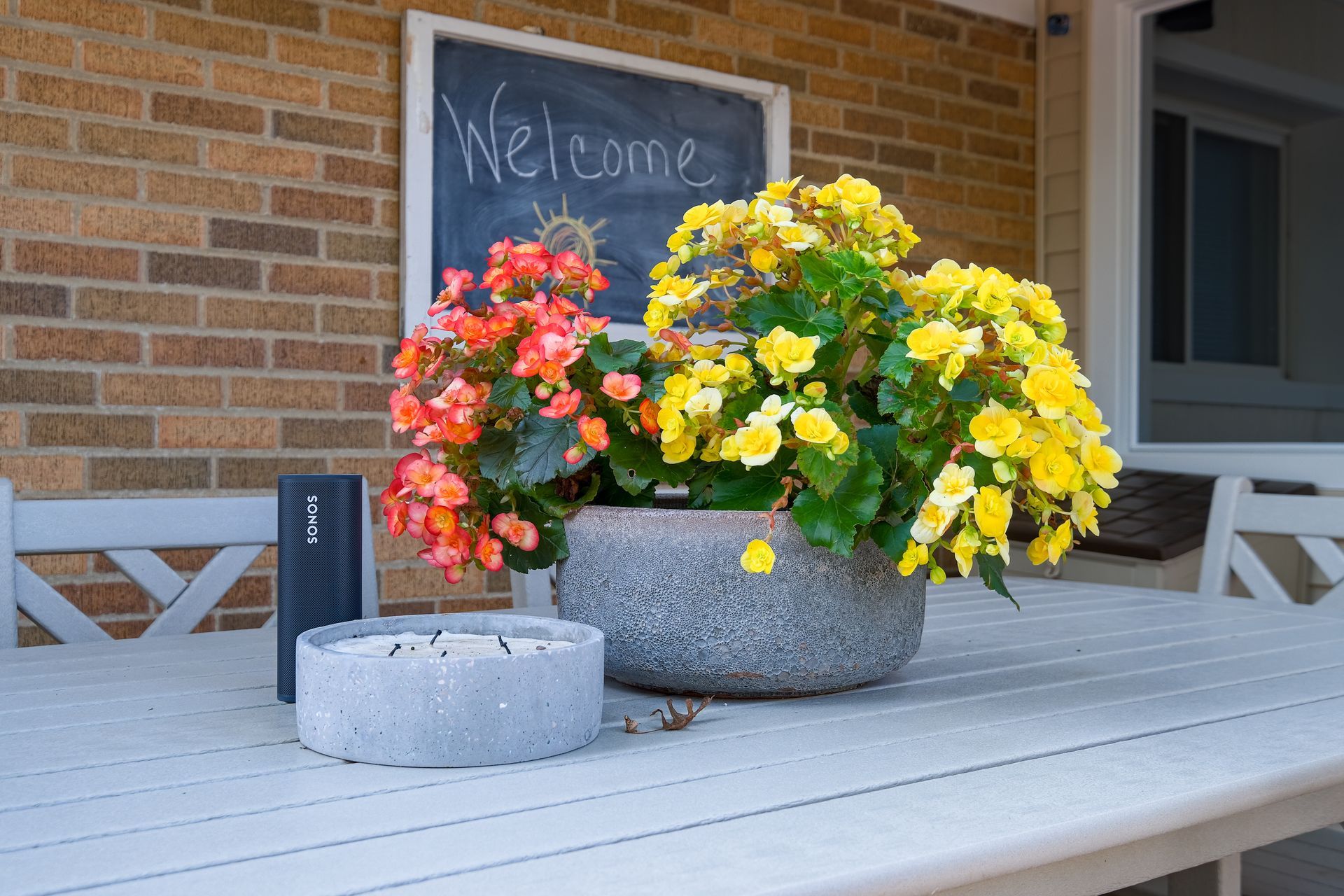 Table with flowers and a welcome sign on a porch. Flowers are orange and yellow, and the sign is on a brick wall.