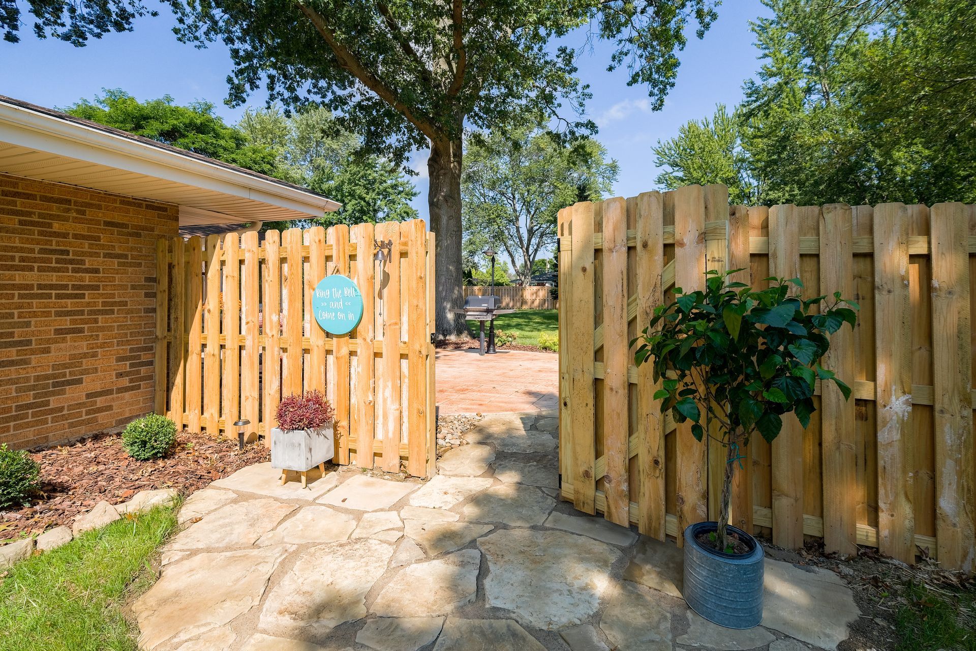 Wooden fence with open gate leading to a brick patio. A potted tree is on the right.