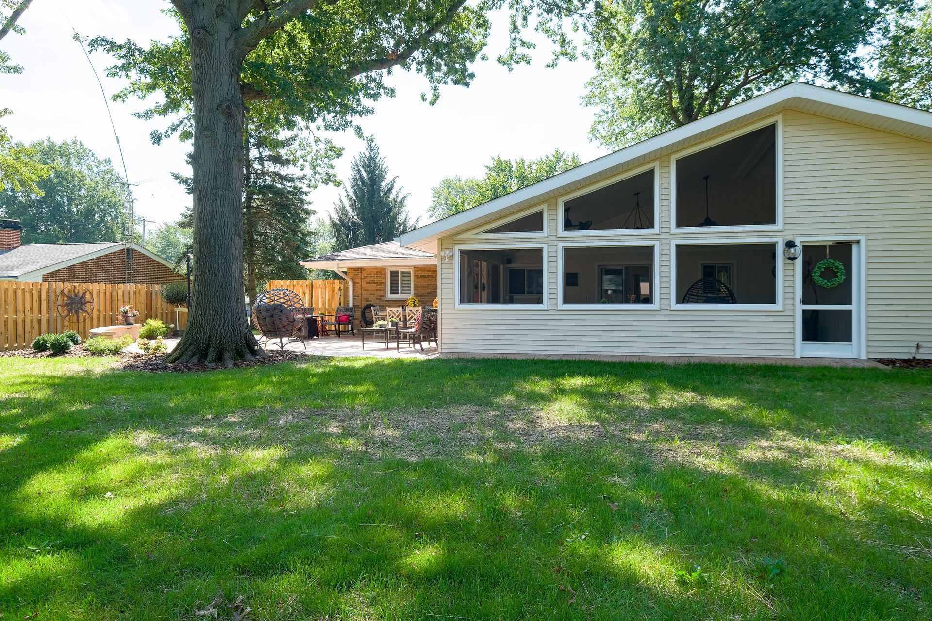 Backyard with a light beige house, large windows, and a patio with chairs; green grass and a large tree in the foreground.