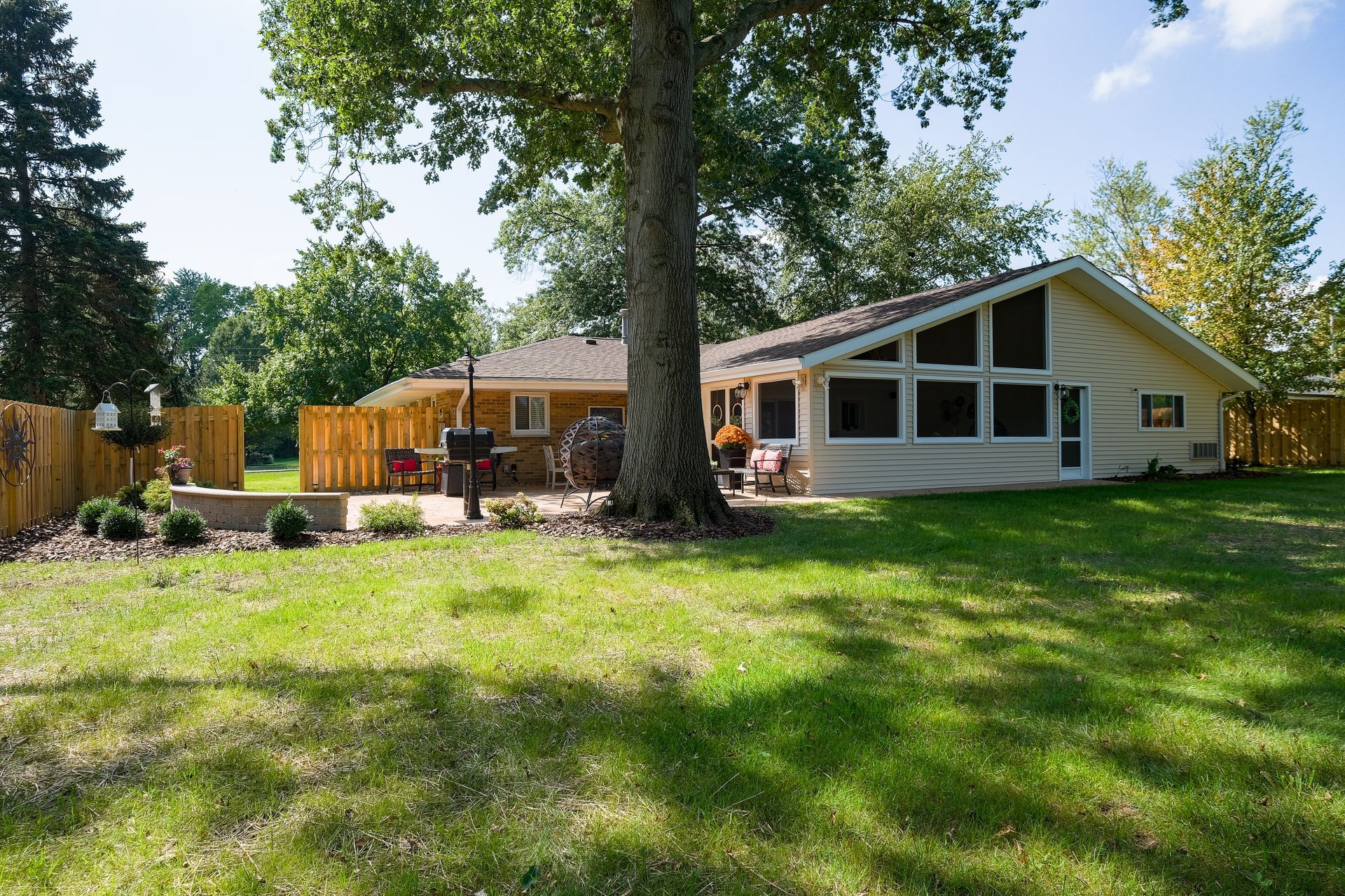 Backyard with house and patio; a tree is center. Green grass, wooden fence, blue sky.