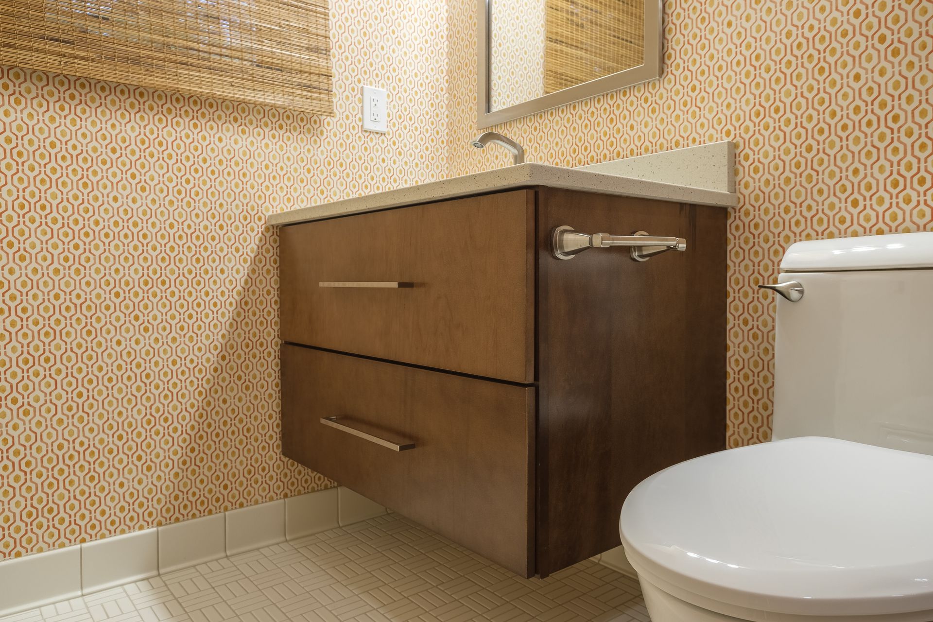 Floating wooden bathroom vanity with two drawers, white toilet, patterned orange wallpaper, and blinds.