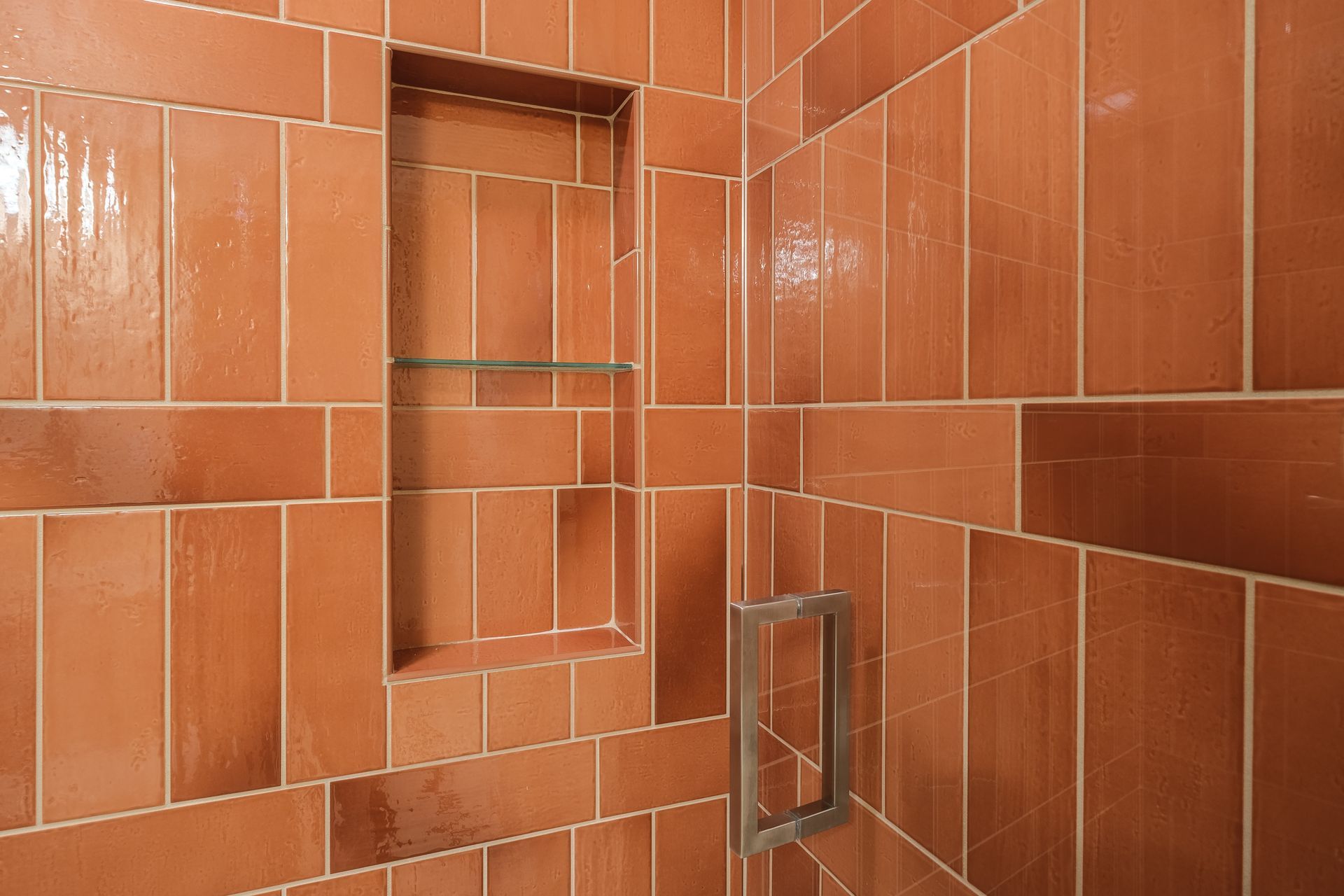 A shower interior with orange rectangular tiles and a recessed shelf, next to a silver door handle.