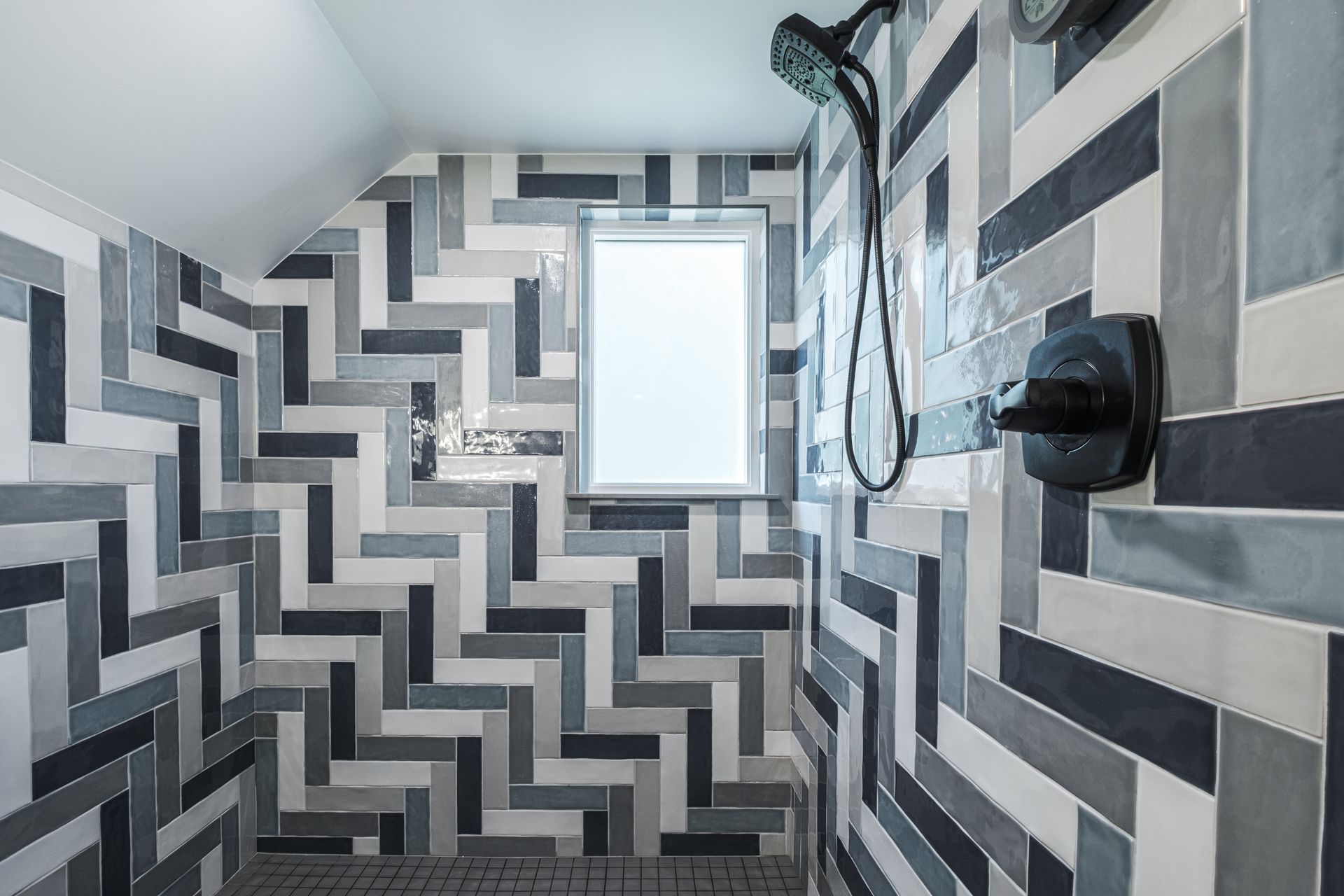 Herringbone-tiled shower in various shades of gray, black, and white. Features a small window and dark shower fixtures.