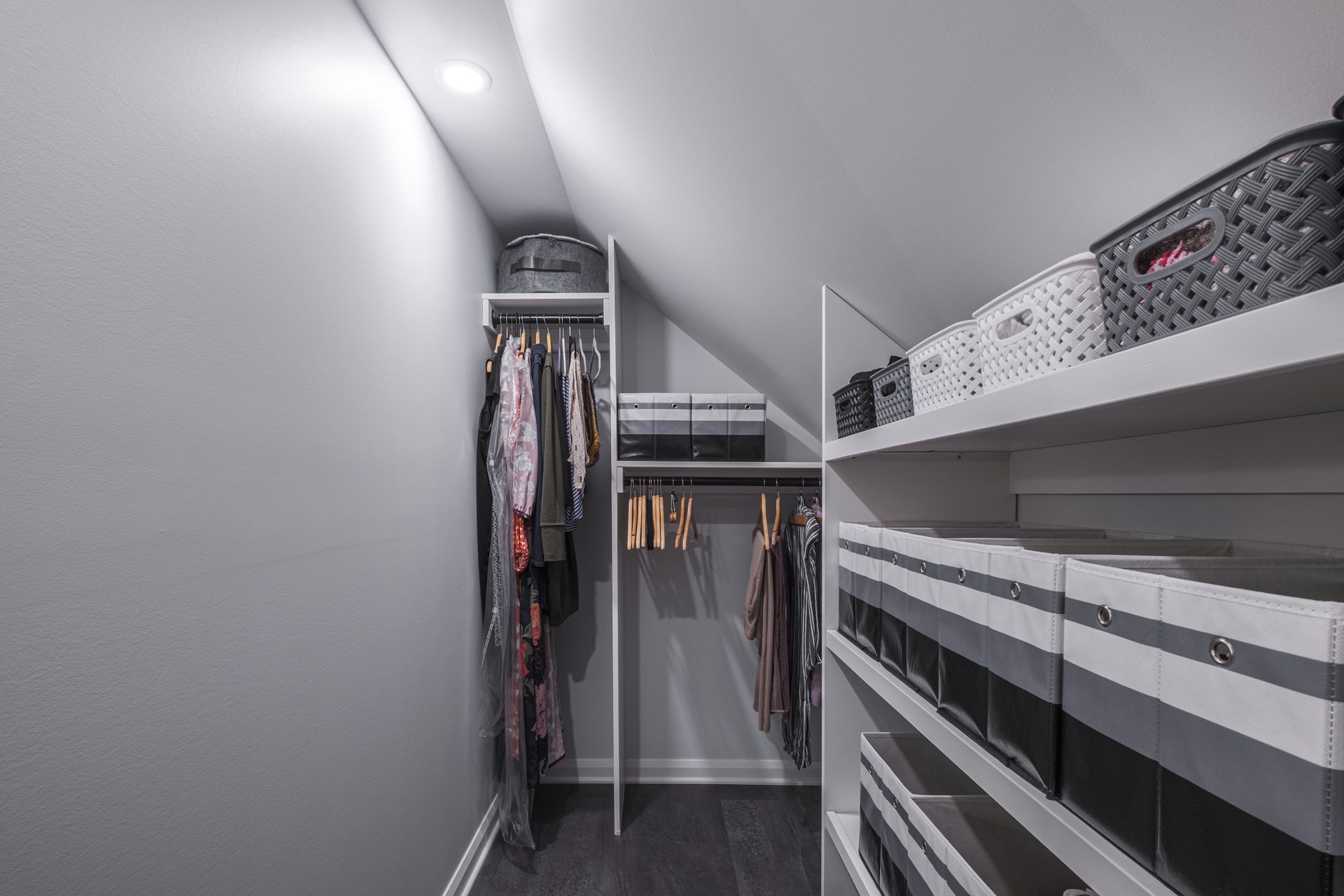 Walk-in closet with sloped ceiling. Gray walls, dark floor. Clothing hangs, shelves hold baskets and storage boxes.