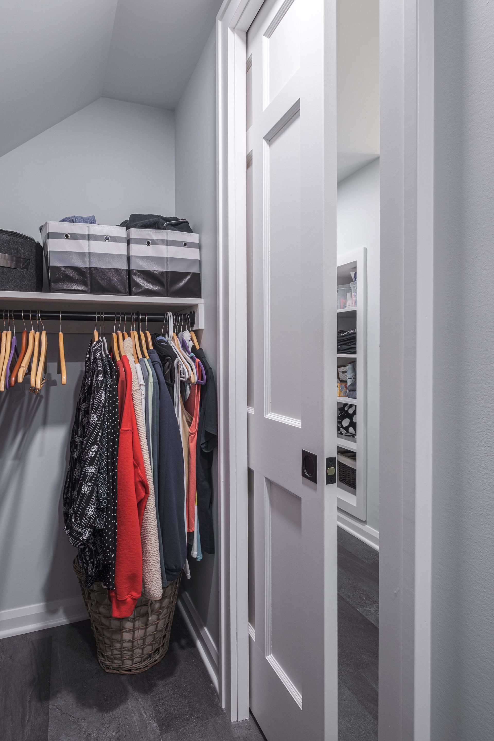 White door open to a walk-in closet with clothes hanging on racks and storage bins above.
