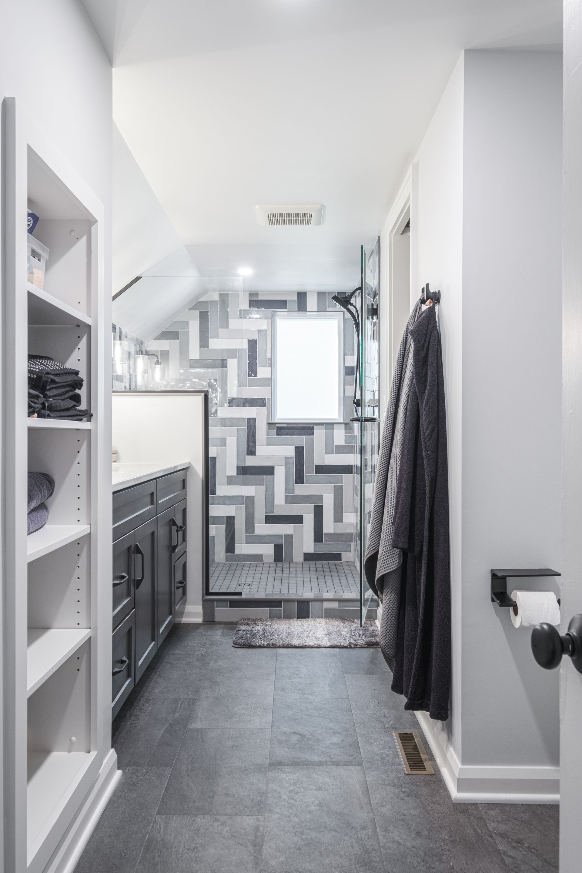 Narrow bathroom with gray tile, shower with geometric tiles, and built-in shelves on left.
