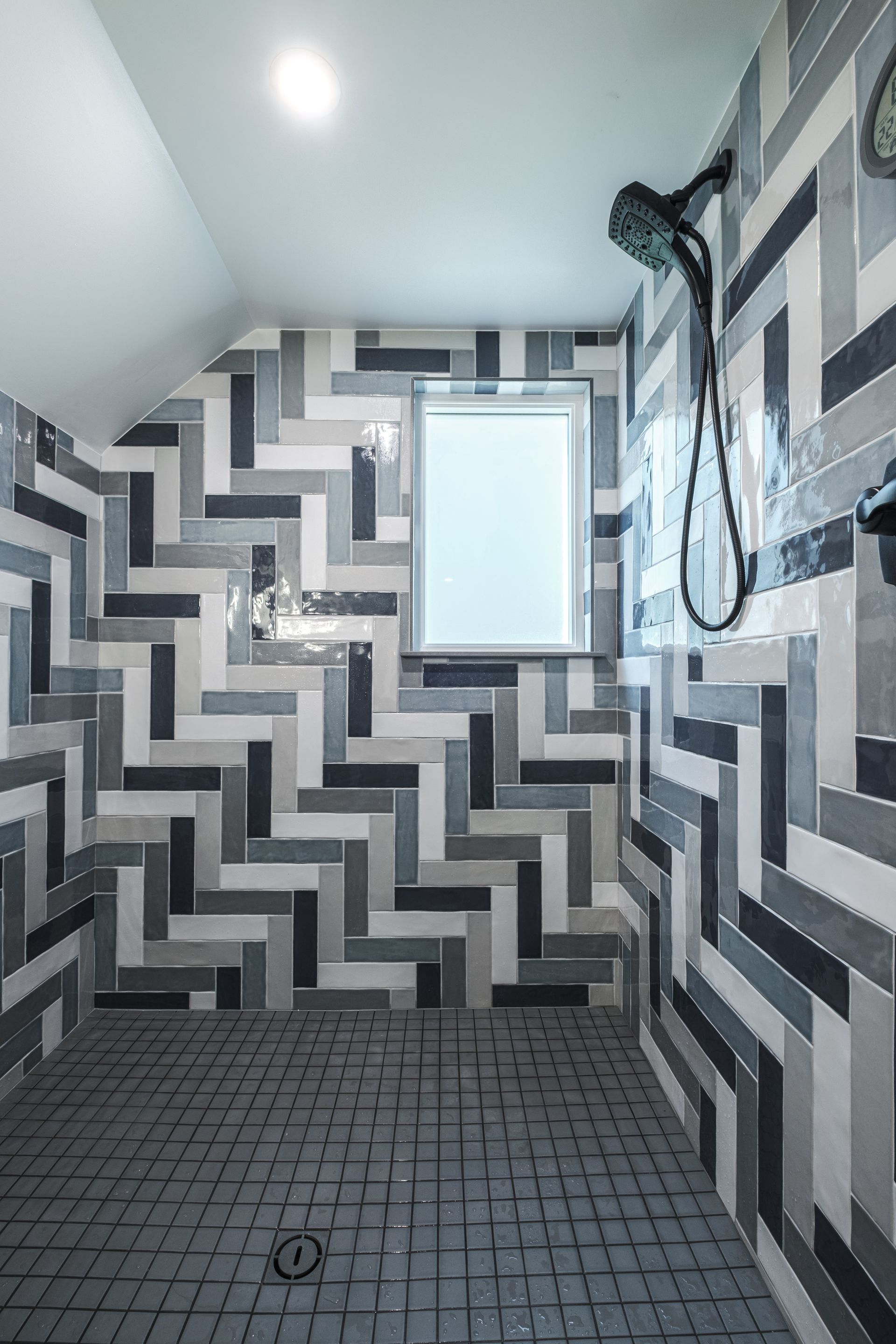 Shower with herringbone pattern tiles in gray and white, showerhead, window.