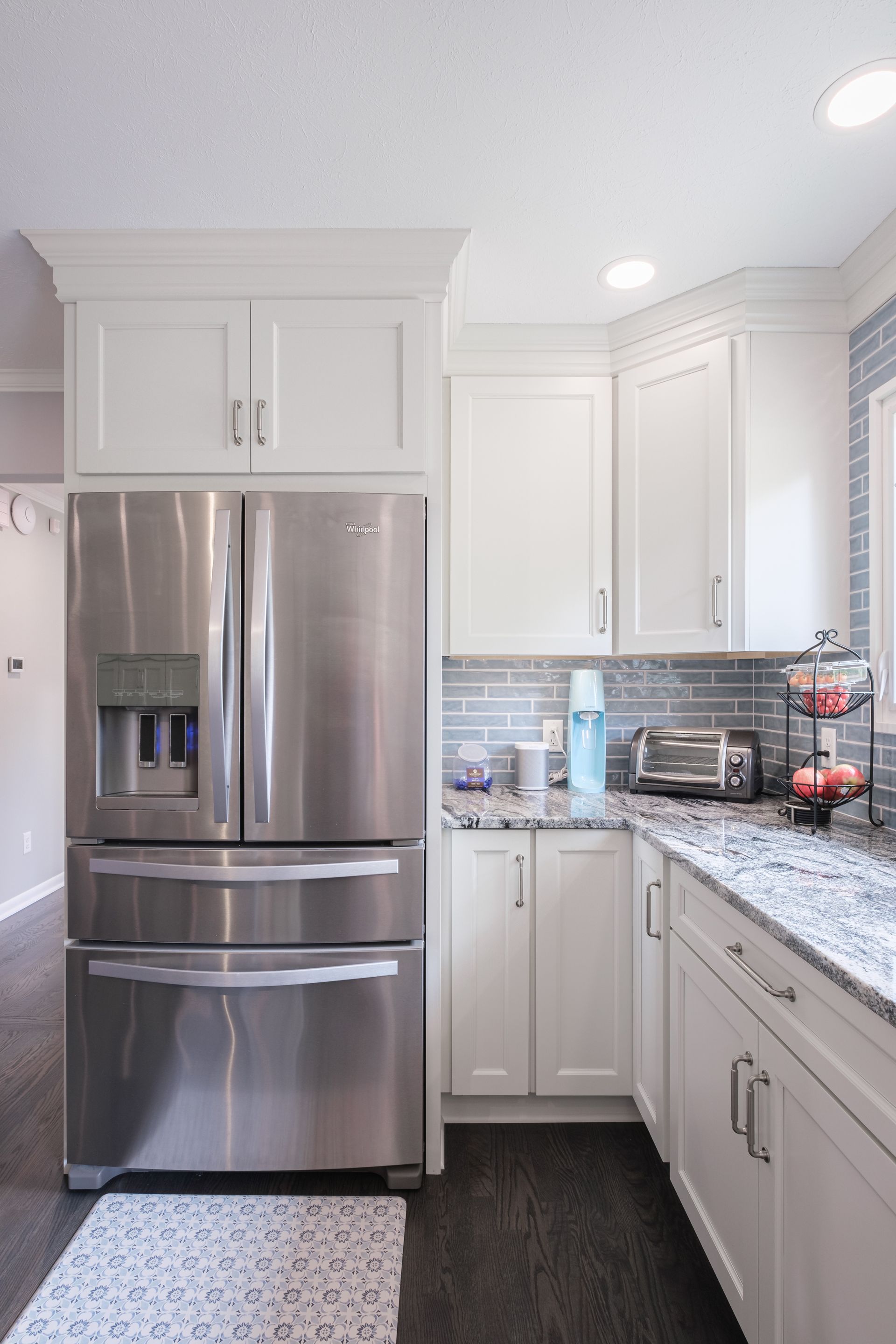 Stainless steel refrigerator and white cabinets in a bright kitchen. Granite countertop, gray backsplash, and dark floor.