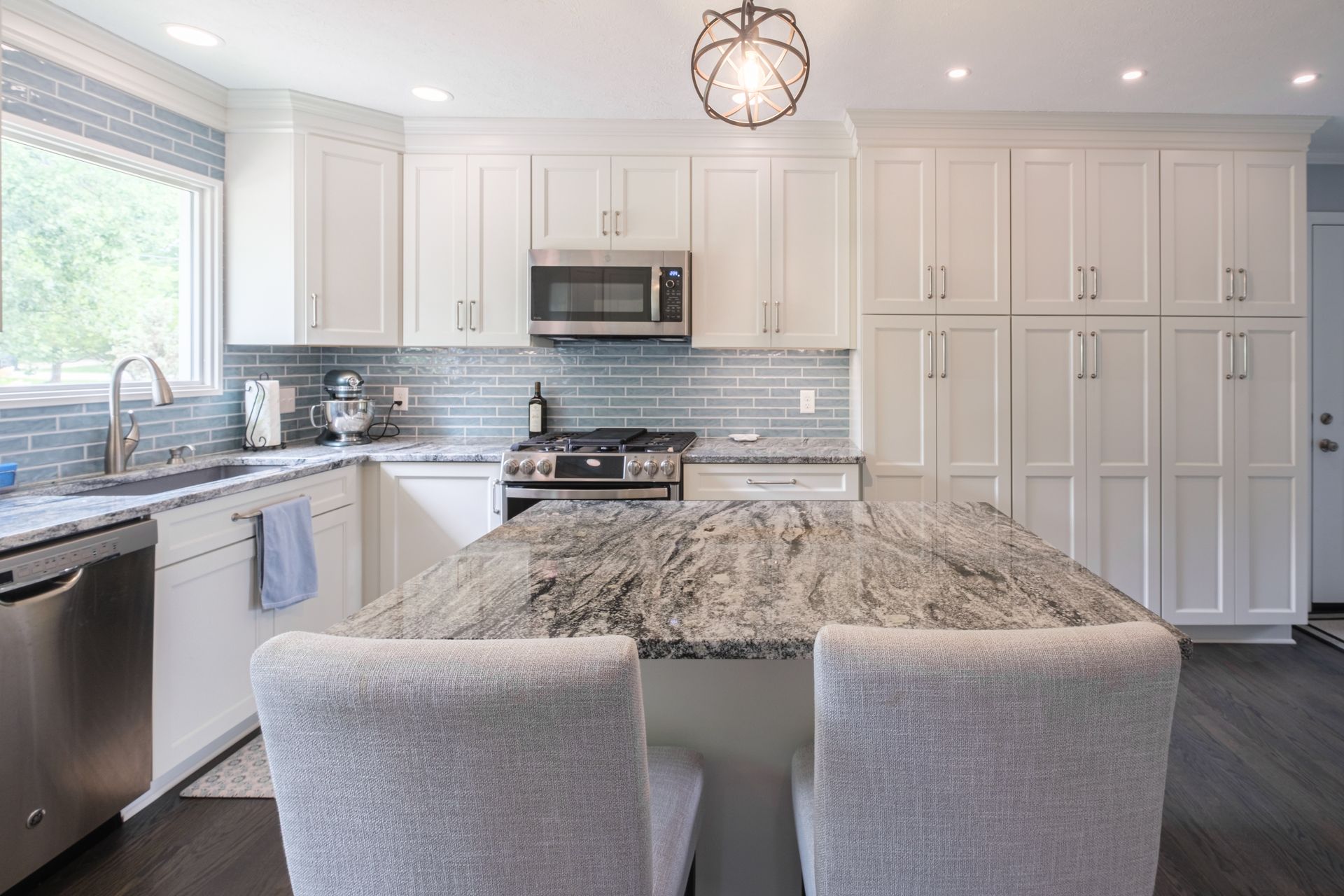 White kitchen with gray island, light blue backsplash, stainless steel appliances, and two gray chairs.