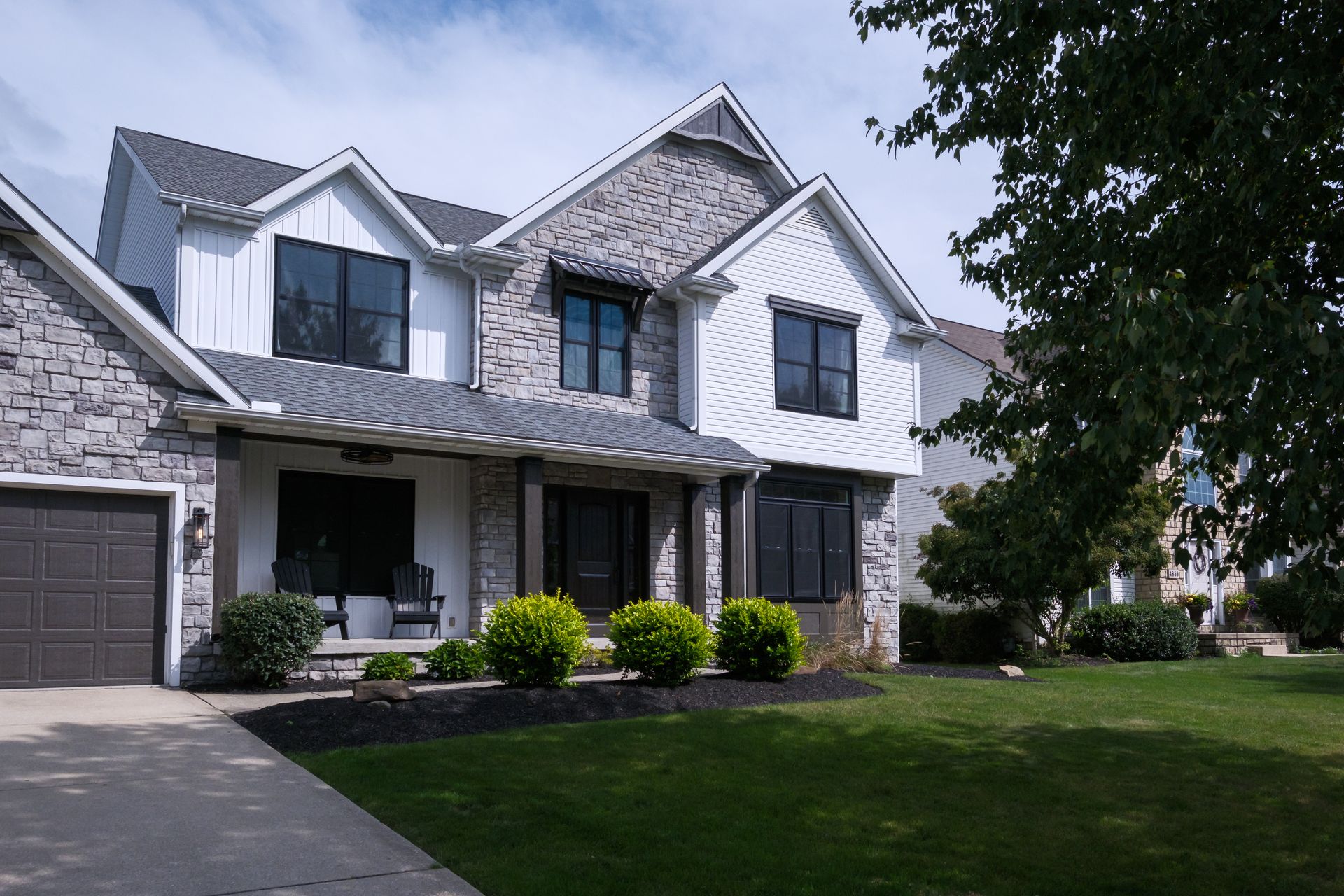 Two-story house with gray stone and white siding, black windows, a garage, and a green lawn.