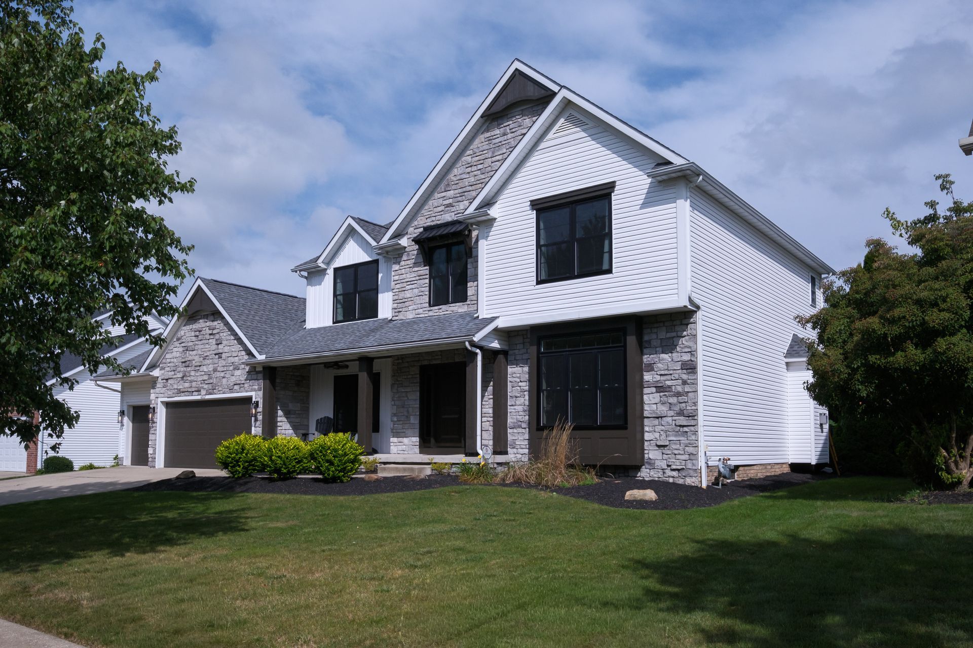 Two-story house with gray stone accents, white siding, black windows, and a green lawn under a partly cloudy sky.