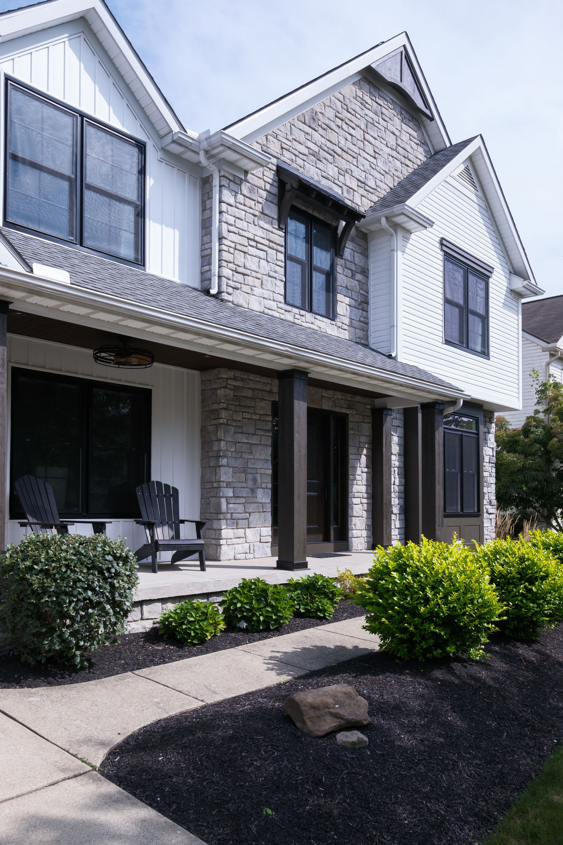 Two-story house with stone and white siding, black windows, porch, and landscaped yard.
