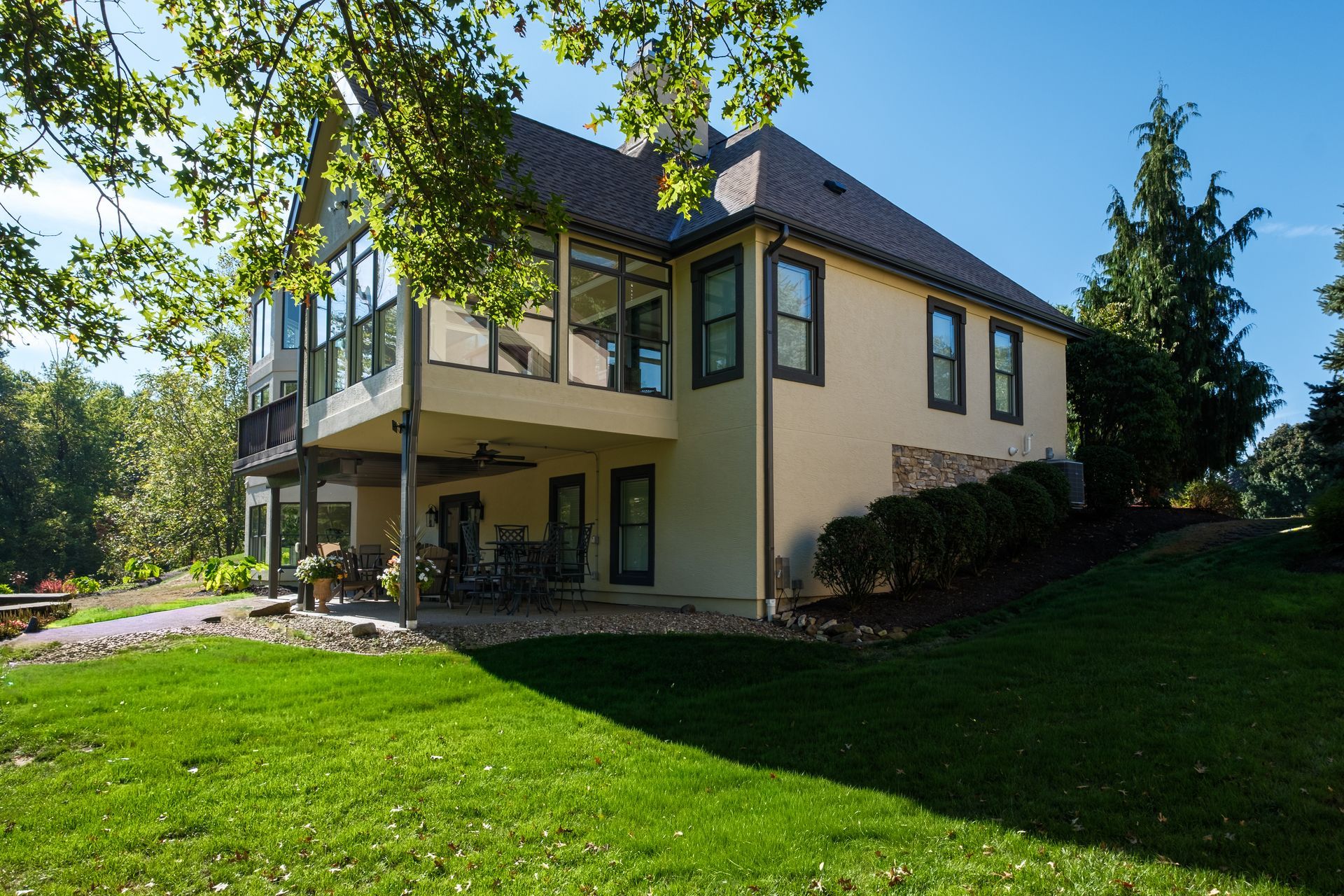 Two-story house with tan stucco exterior, large windows, and a covered lower level, set on a grassy hill.