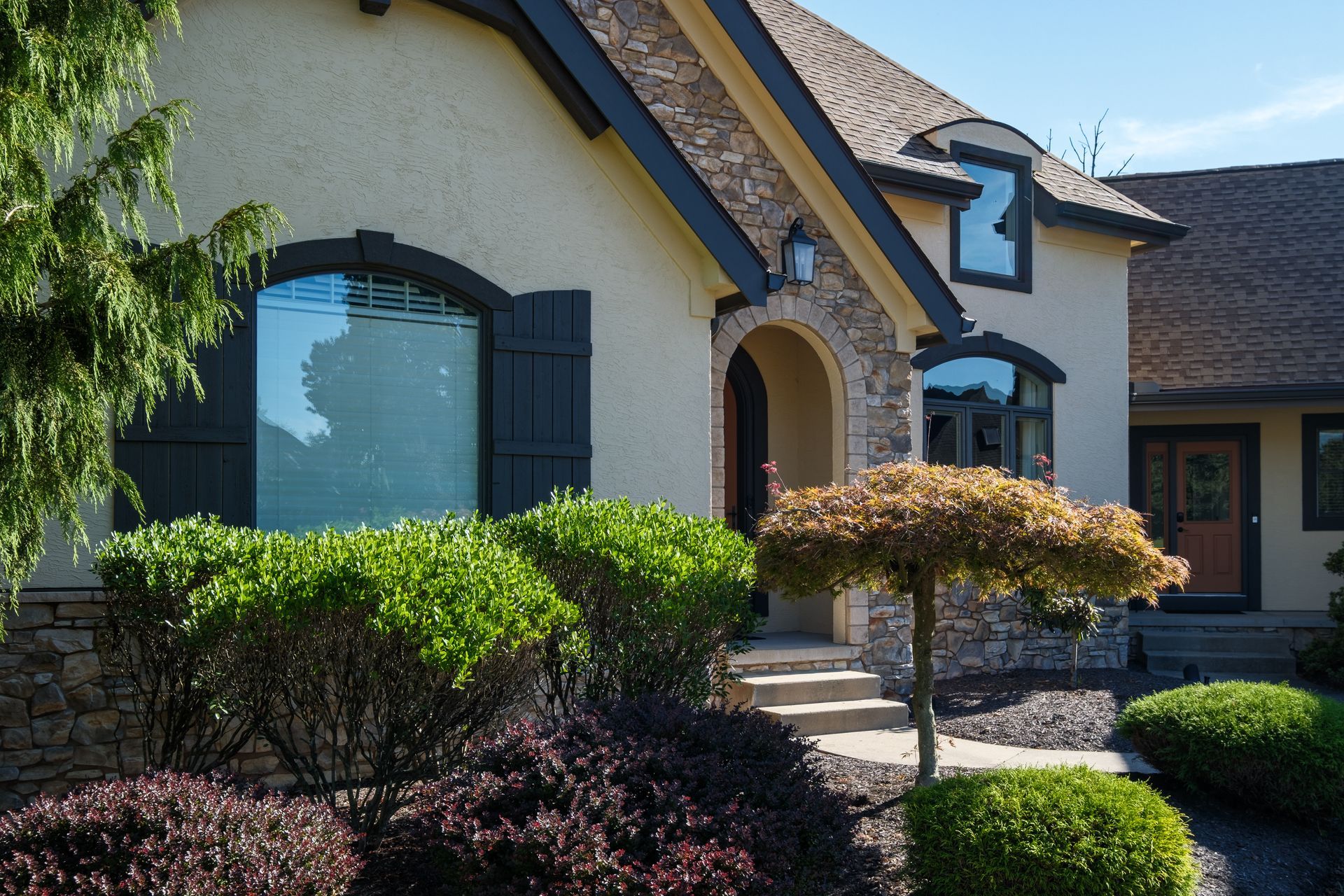 Tan stucco house with dark trim, stone accents, and landscaped front yard with shrubs and small tree.