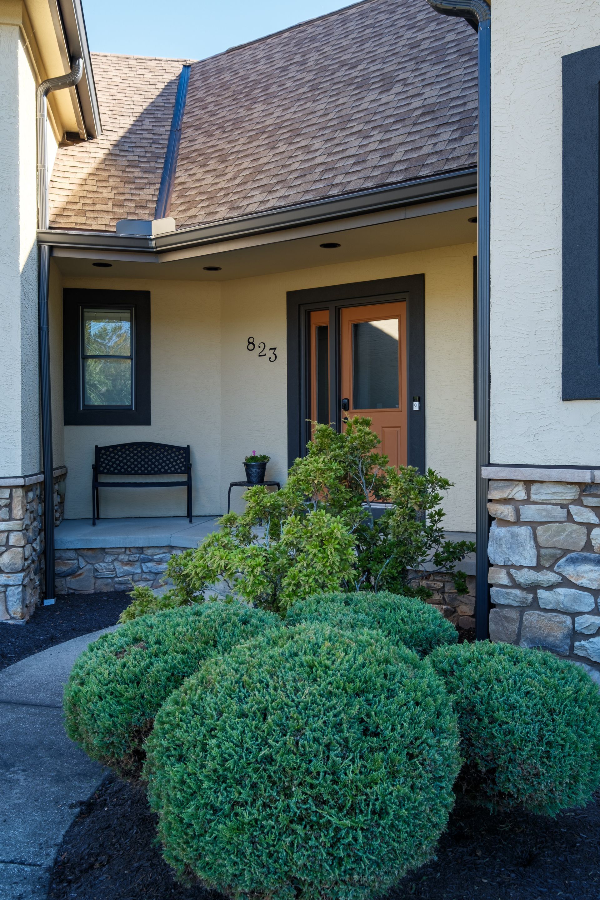 Front entrance of a house with rounded green bushes, a bench, and a brown door; the house number is 553.