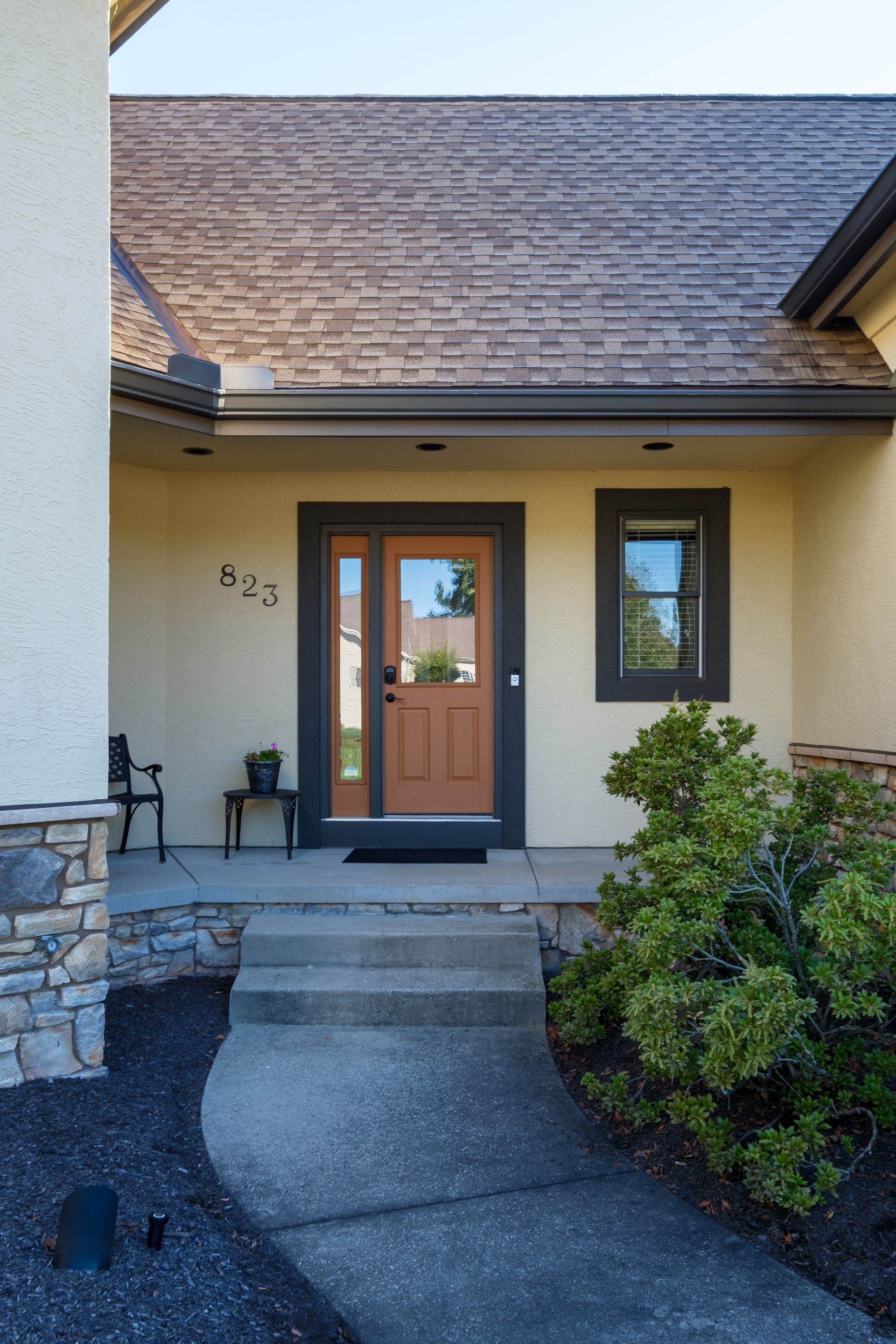 Beige house entrance with steps leading to a brown door, flanked by a small window and a shrub.