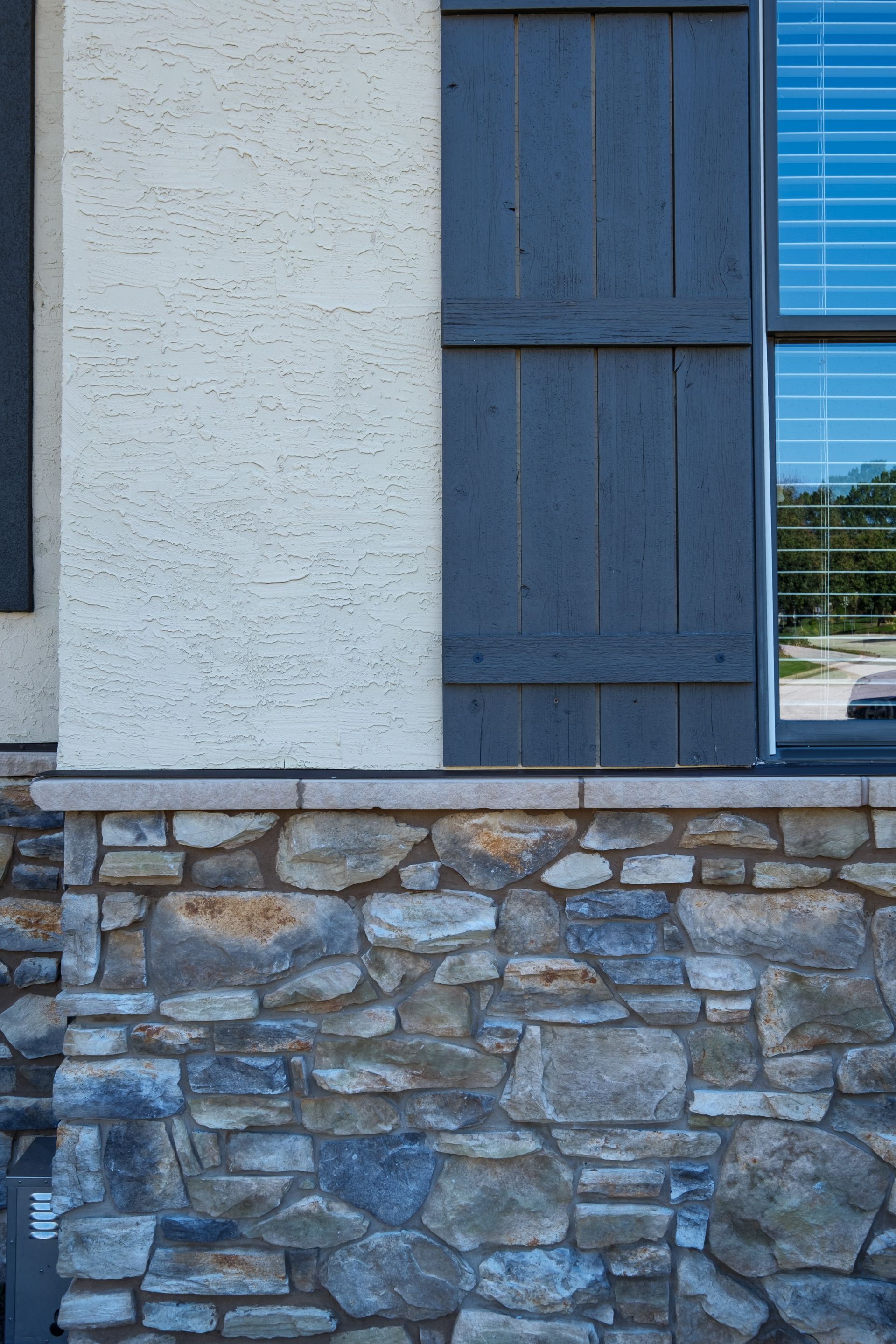 Exterior of a building with stone facade, light stucco, blue shutters, and a window reflecting sky.