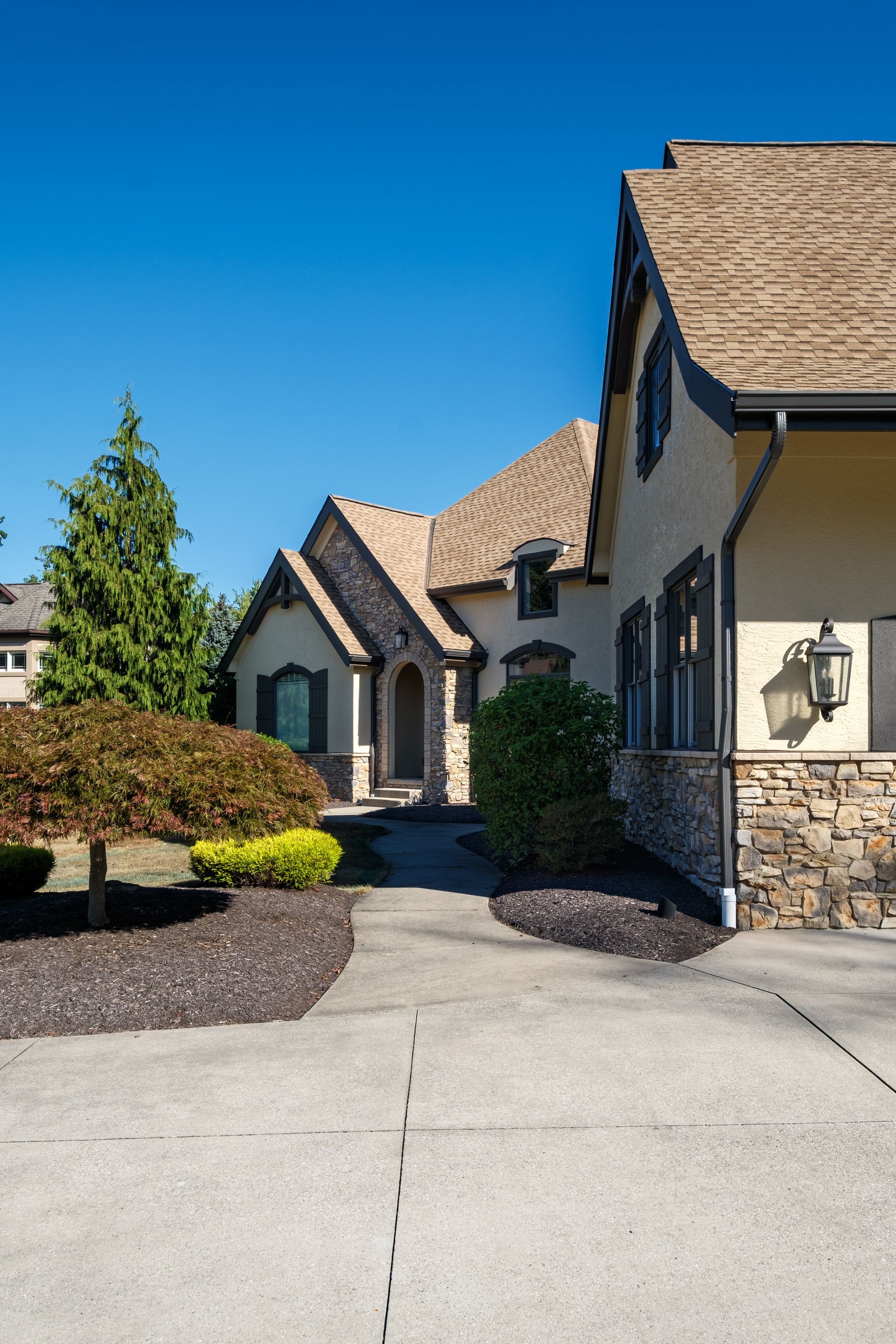A house with a beige facade, brown roof, and stone accents, with a driveway and landscaping under a blue sky.