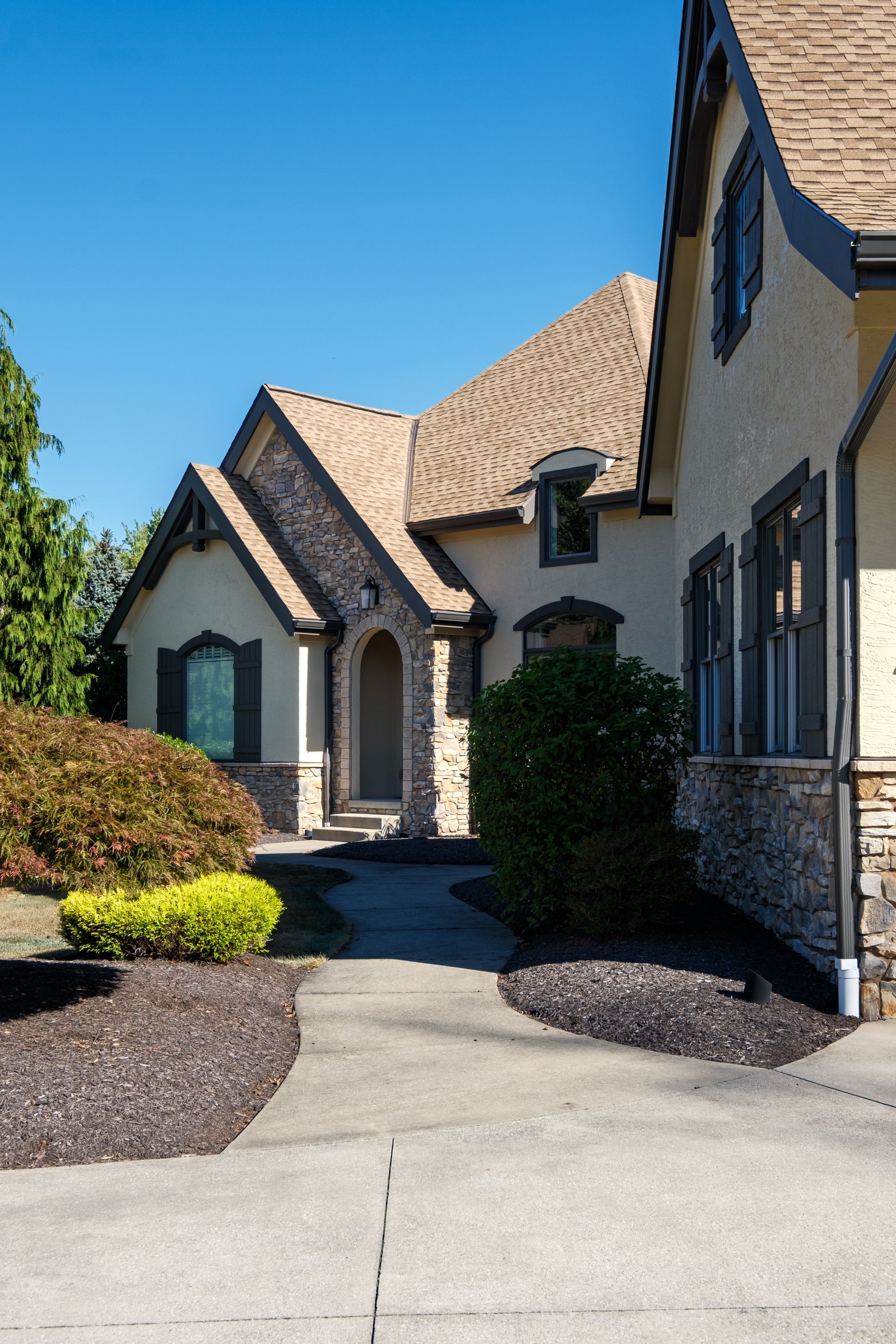 Tan stucco house with stone accents, arched entry, and brown roof. Paved walkway leads up to the front door.