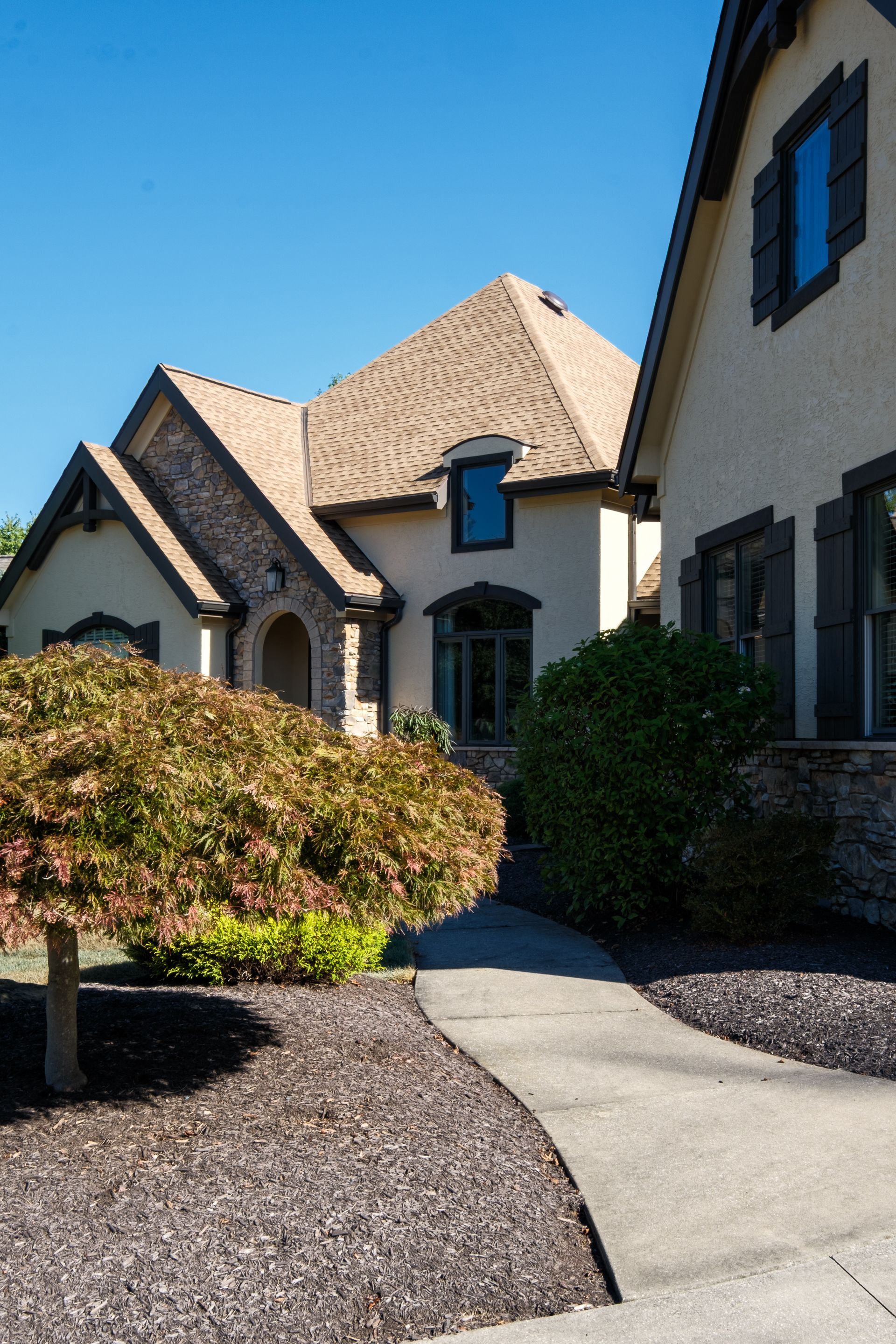 A winding walkway leads to a beige home with a brown roof and stone accents on a sunny day.