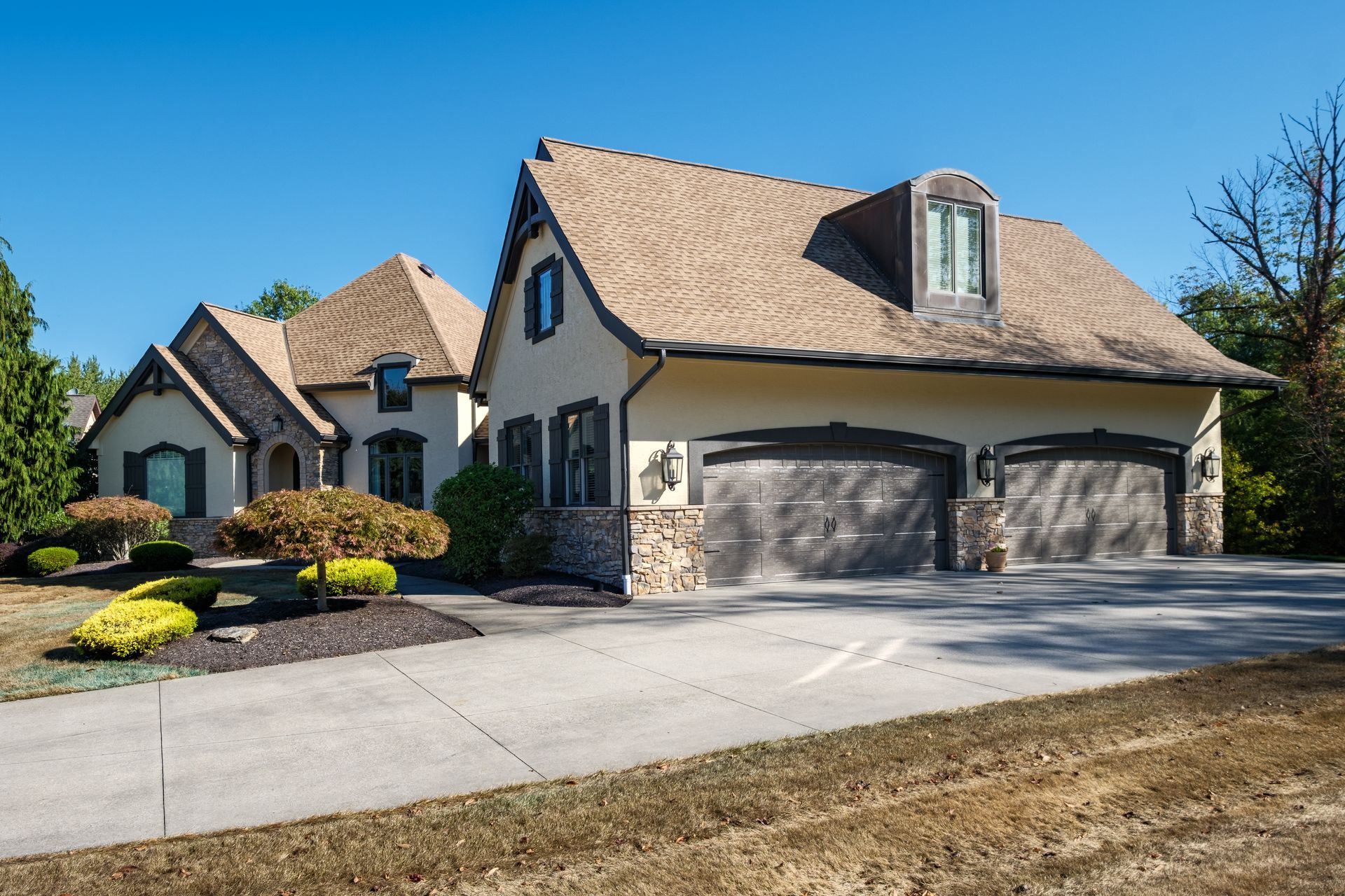 Tan stucco and stone house with a two-car garage; driveway, landscaping, and blue sky.