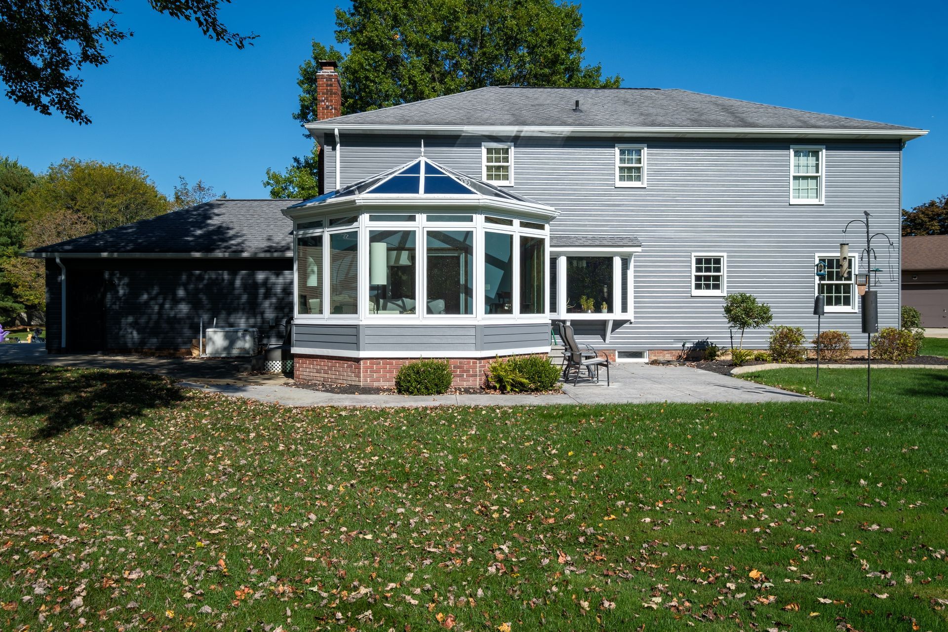 Backyard view of a two-story gray house with an attached sunroom, brick base, and lush lawn on a sunny day.