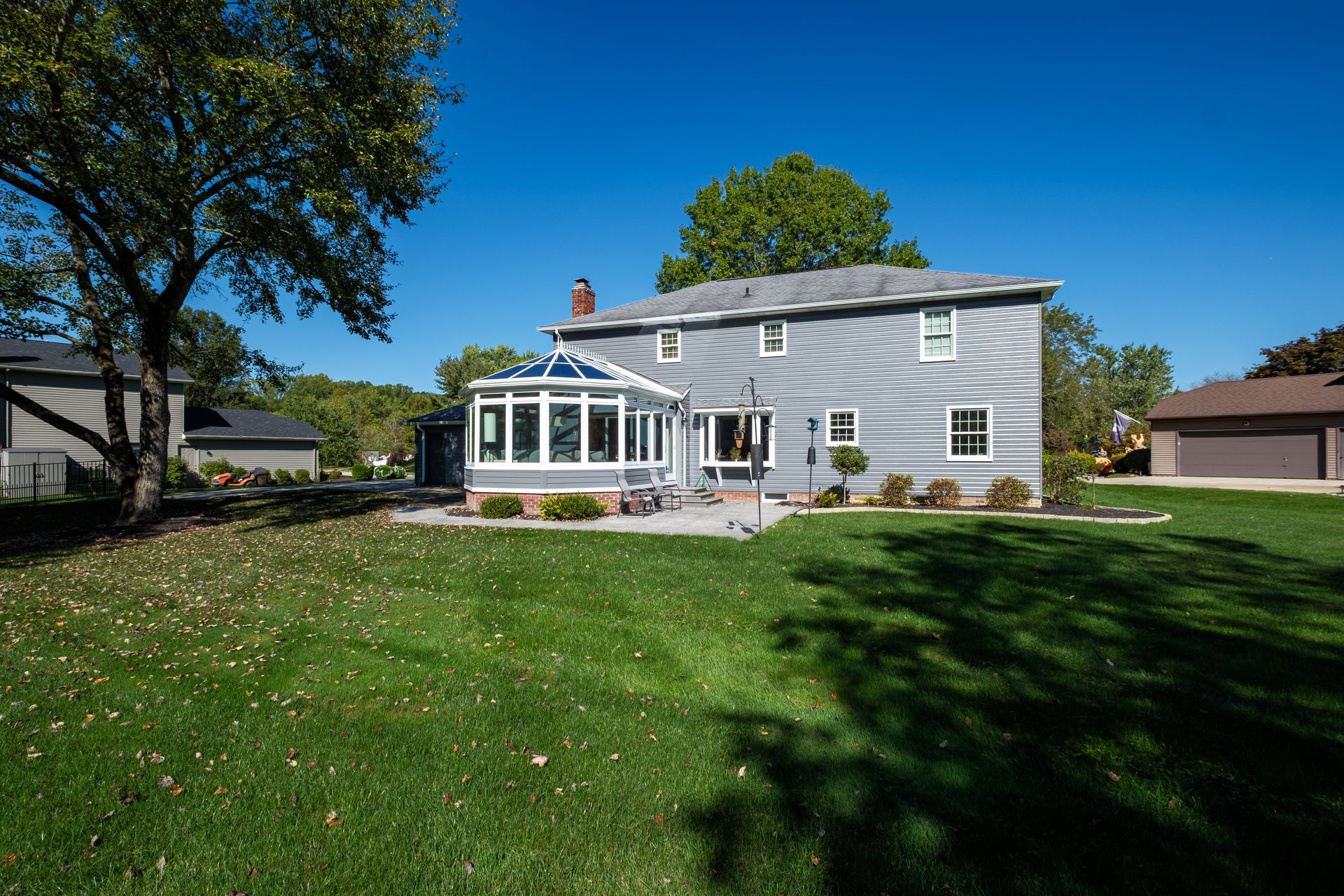 Backyard of a two-story gray house with a sunroom, patio, and green lawn on a sunny day.