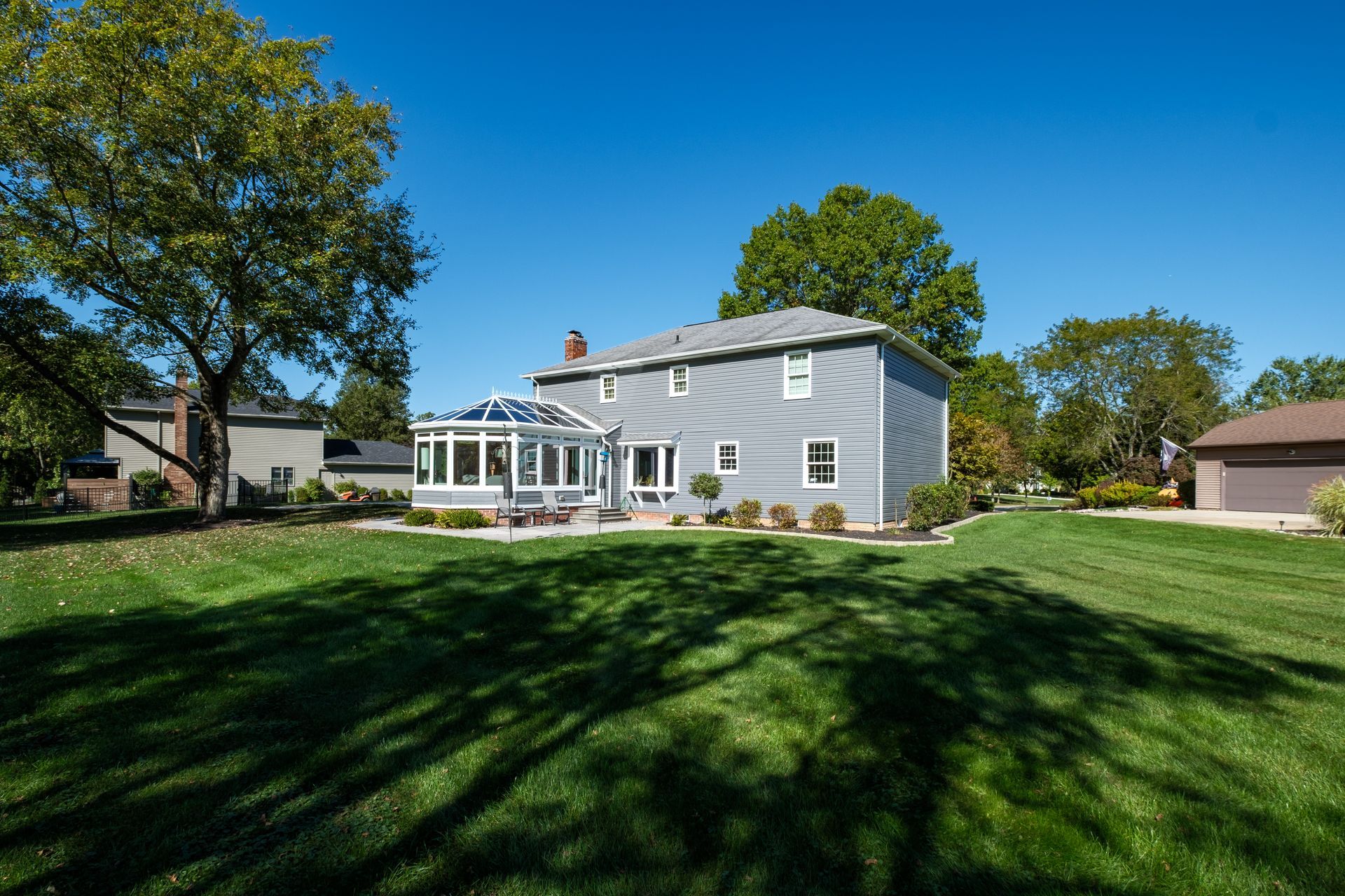 Two-story gray house with sunroom, green lawn, trees, and blue sky.