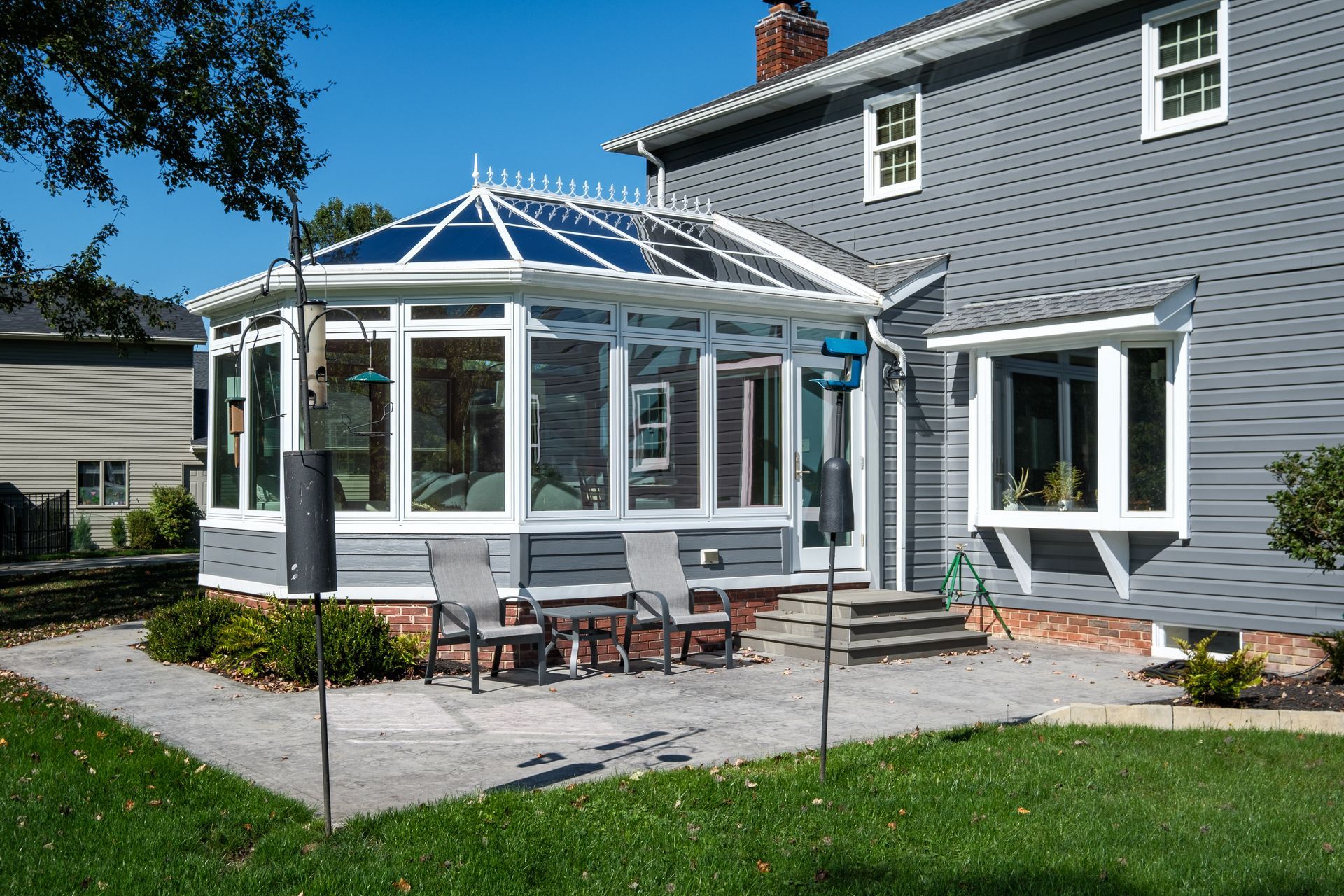 A sunroom with a glass roof and walls, attached to a gray house. Chairs and a patio are in the yard.