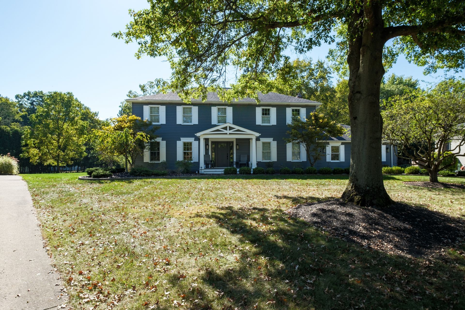 Two-story blue house with white trim and shutters, surrounded by a lawn and trees on a sunny day.