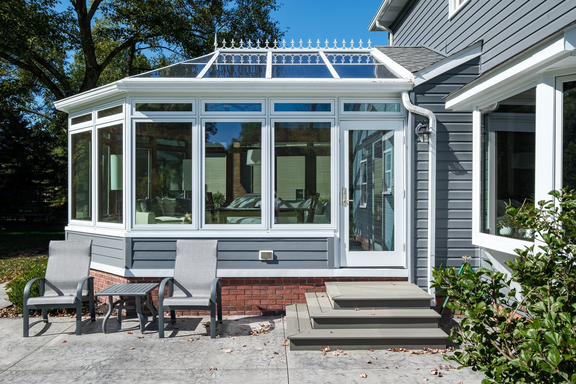 Sunroom addition to a gray house, with seating, glass windows, and a brick base.