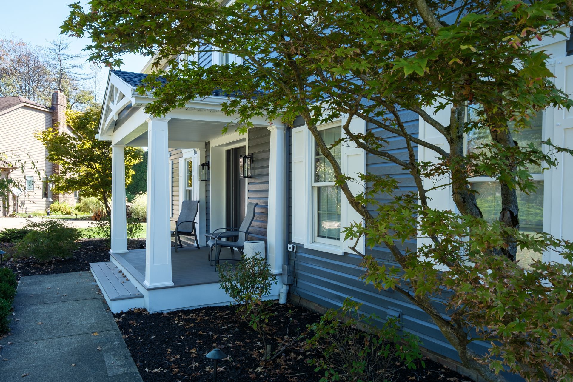 Blue house with white porch, two chairs, and tree in front.